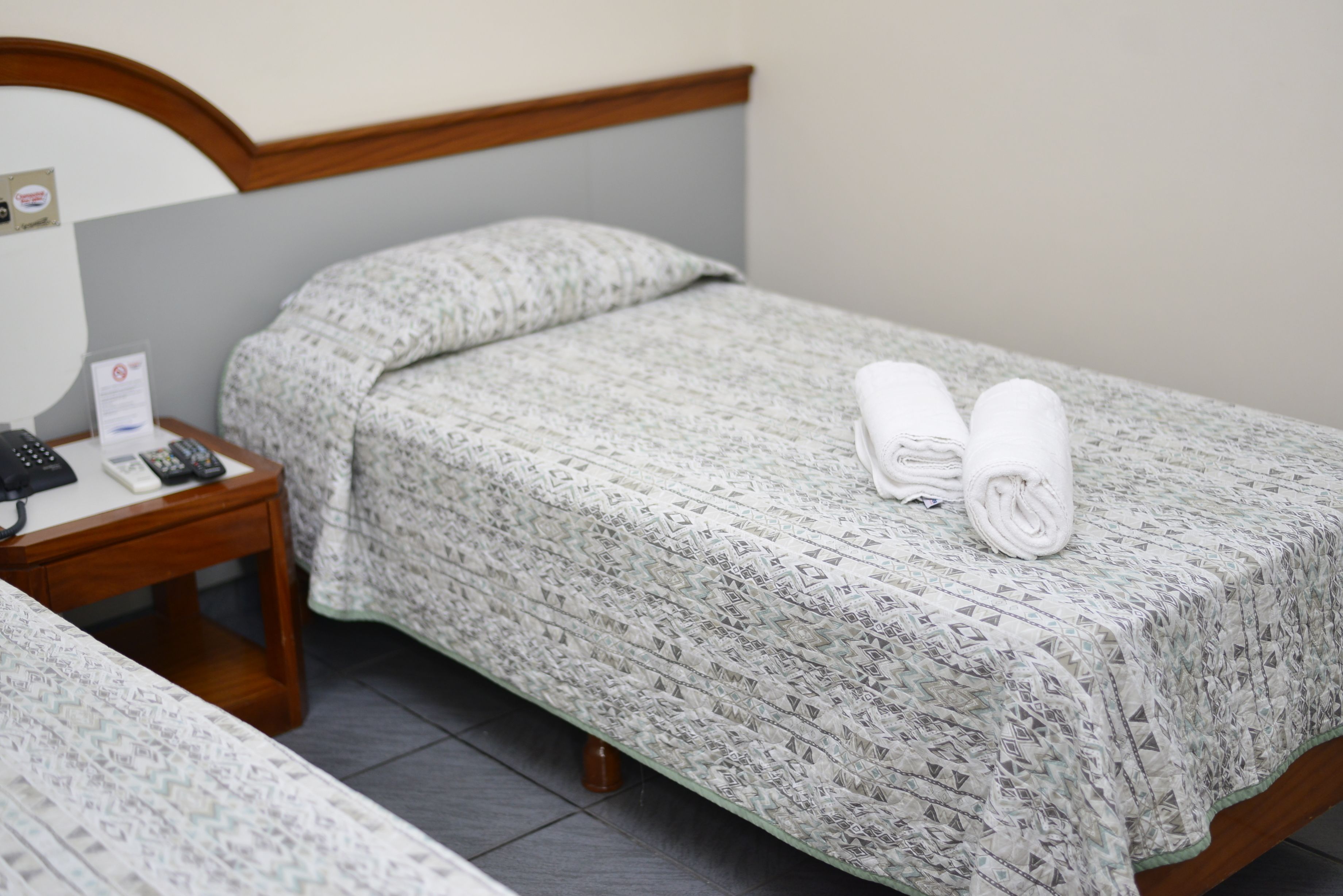 A neatly made bed with a patterned bedspread and two rolled towels. A bedside table with a remote control and a sign is visible.