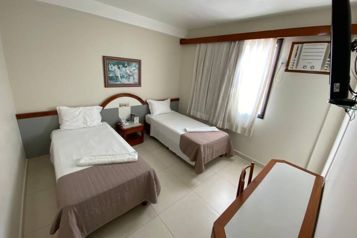 A hotel room with two single beds, a wooden nightstand, a television, and a window with curtains.