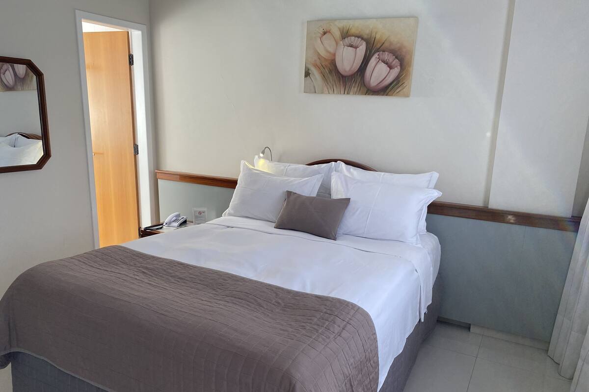 A neatly made bed with white and brown bedding, a bedside phone, and a painting of flowers on the wall.