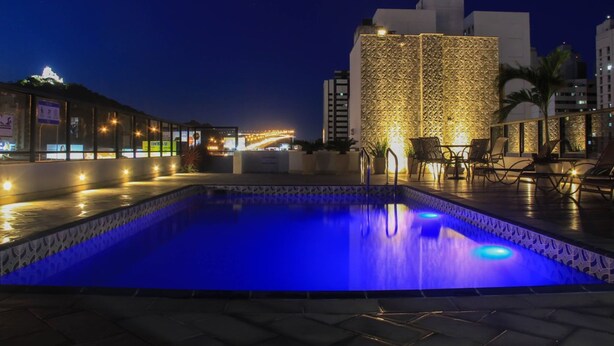 Zona de piscina nocturna con piscina iluminada en azul, sillas de descanso y un edificio con diseño en el fondo.