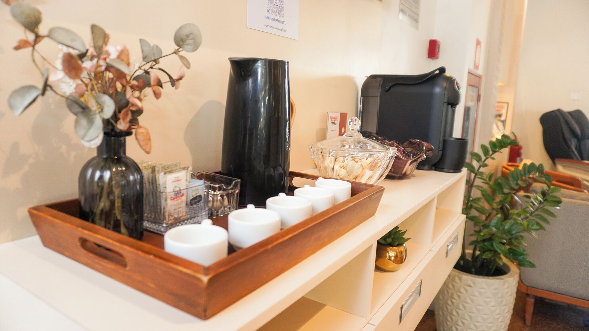 A reception area with a wooden tray holding various items, a vase with dried flowers, a black pitcher, and a coffee machine.
