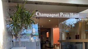 La entrada del Hotel Champagnat Praia, con fachada de vidrio, plantas de interior y piso de baldosas.
