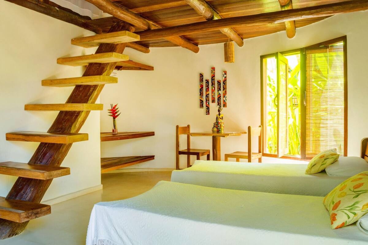 Ein Zimmer mit einer Wendeltreppe aus Holz, zwei Betten, einem kleinen Tisch mit Stühlen und einem Fenster mit Jalousien.