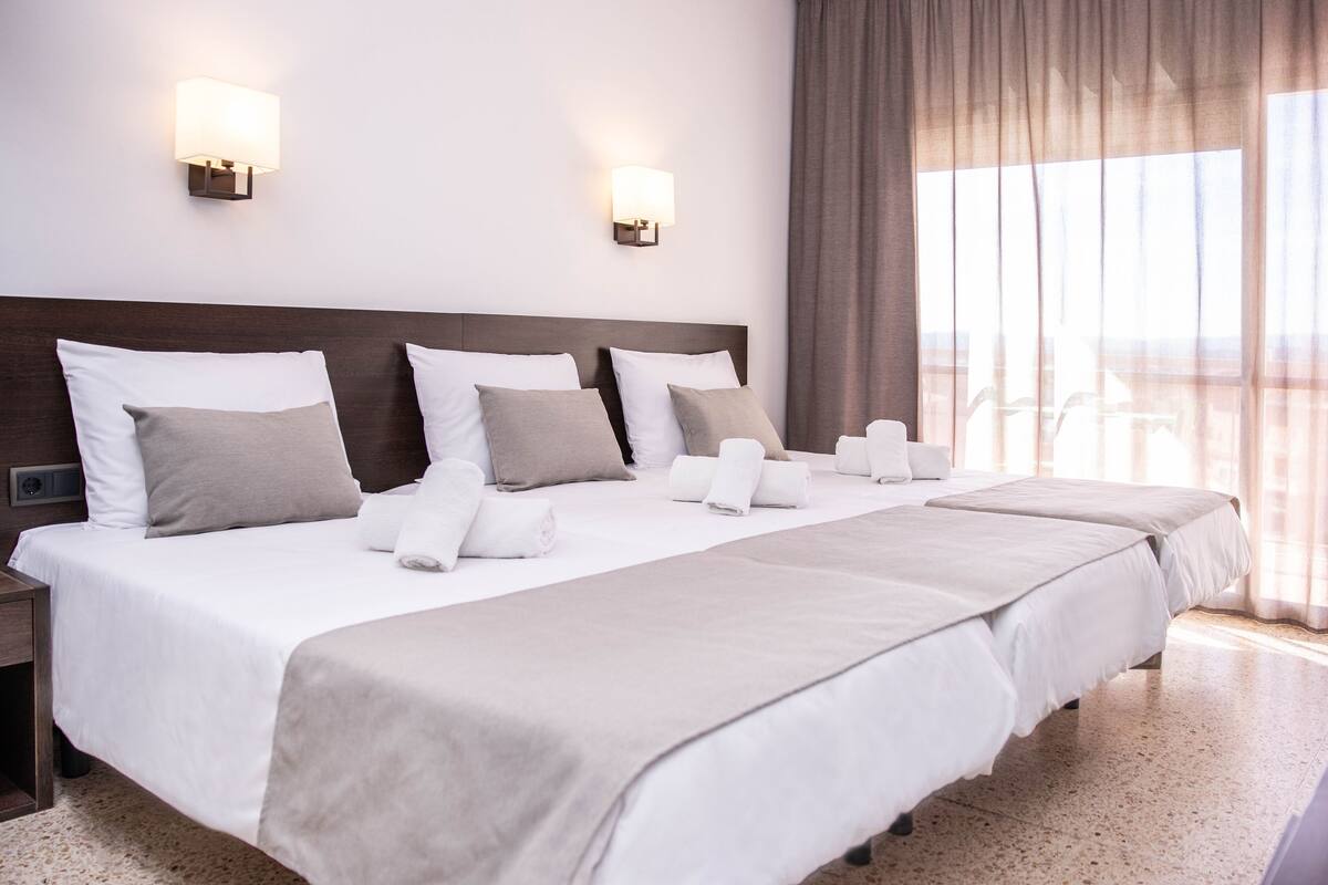 A hotel room with a large bed, two wall-mounted lights, a view of the outdoors, and a window with curtains.