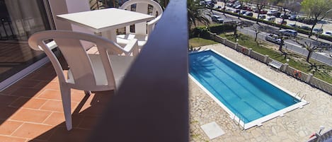 A balcony with a white table and chairs, a pool, and a view of a parking lot and buildings.