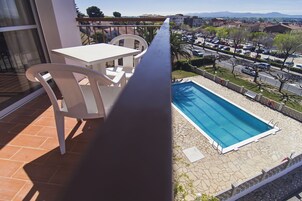 A balcony with a white table and chairs, a pool, and a view of a parking lot and buildings.