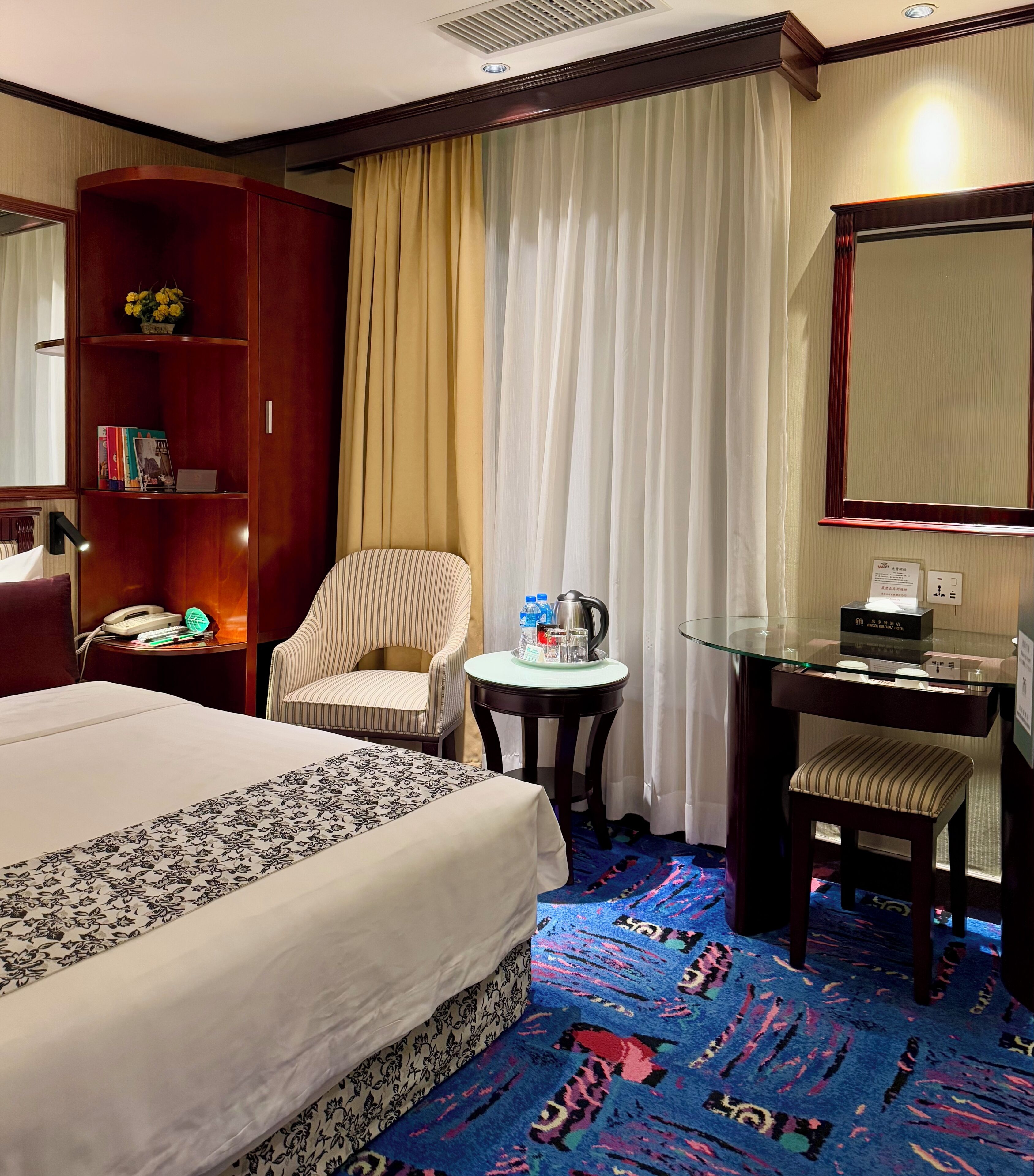 A hotel room with a bed, a chair, a desk, a bookshelf, and a mirror.