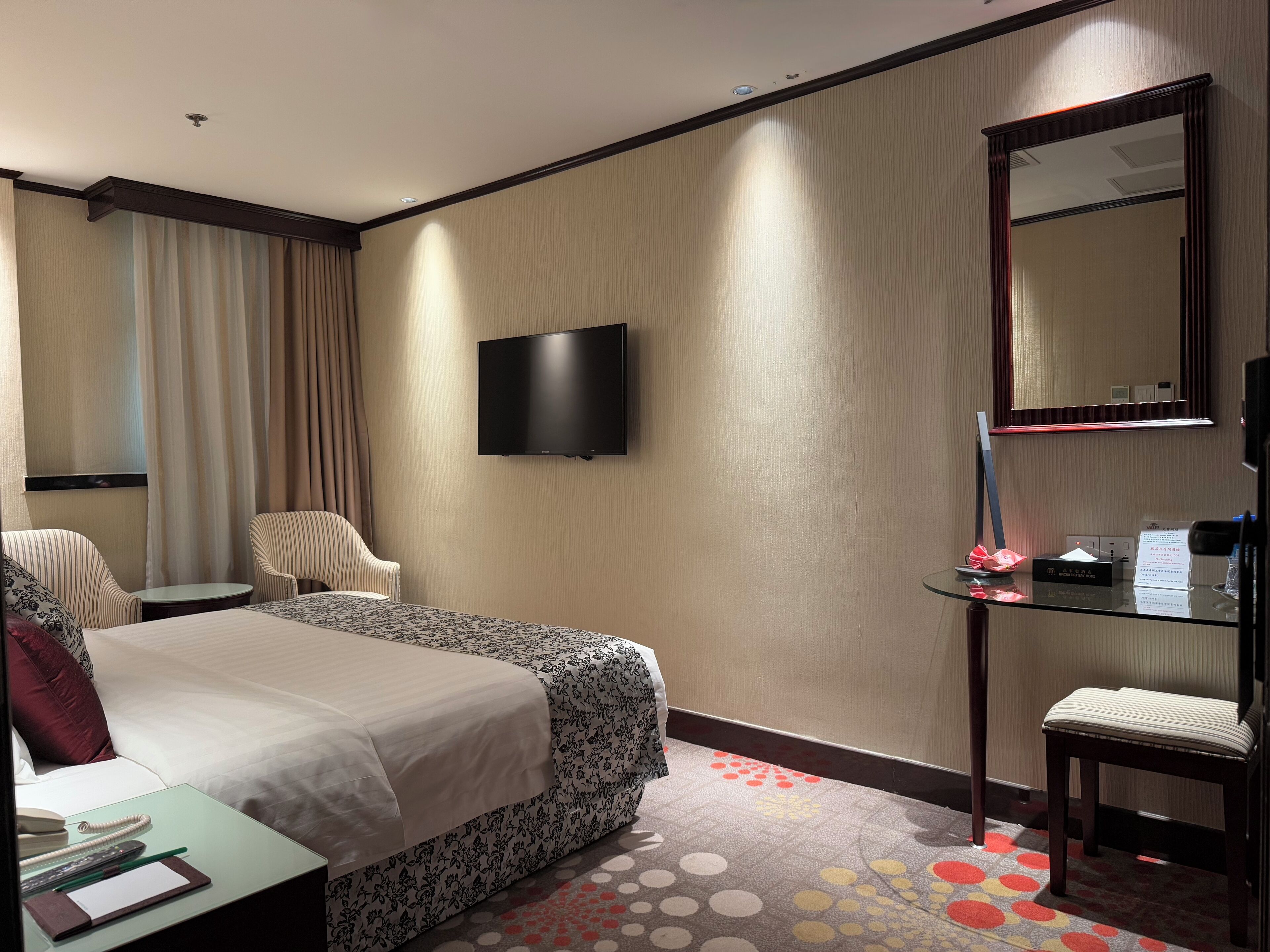 Hotelkamer met een bed, bureau met telefoon en notitieboekje, stoel, televisie aan de muur en een tafeltje met waterfles en menukaart.