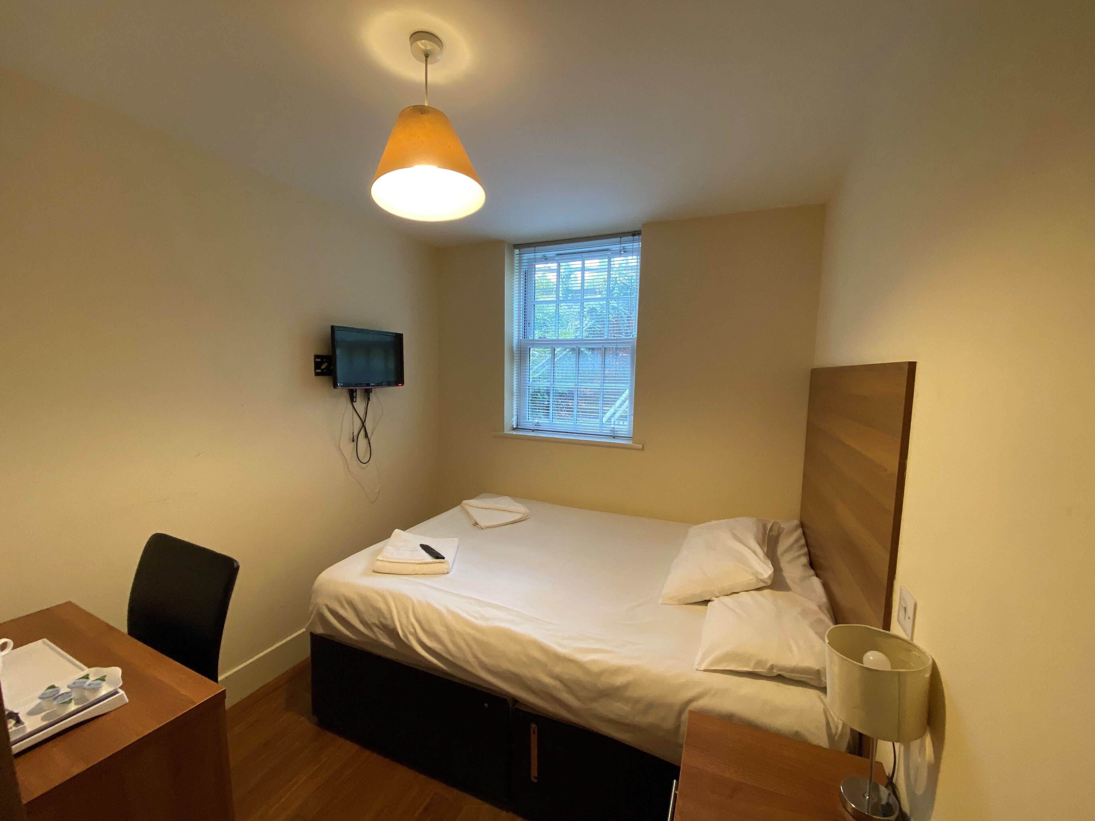 Une petite chambre à coucher, bien rangée, avec un lit simple, un bureau et une chaise, un téléviseur fixé au mur et une fenêtre laissant entrer la lumière naturelle.