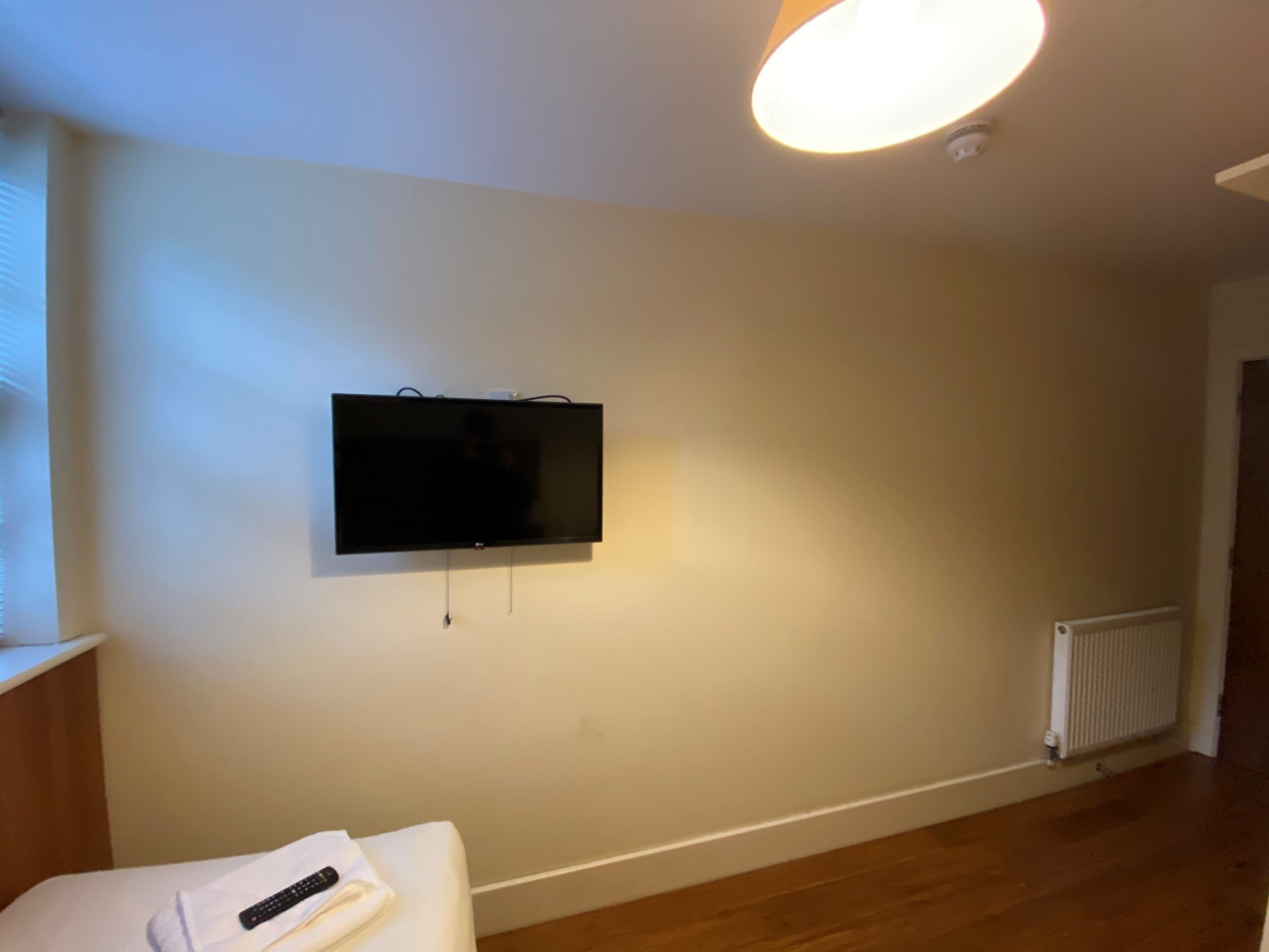 Une chambre avec un téléviseur fixé au mur, un lit blanc avec une télécommande, un plancher en bois et un radiateur.