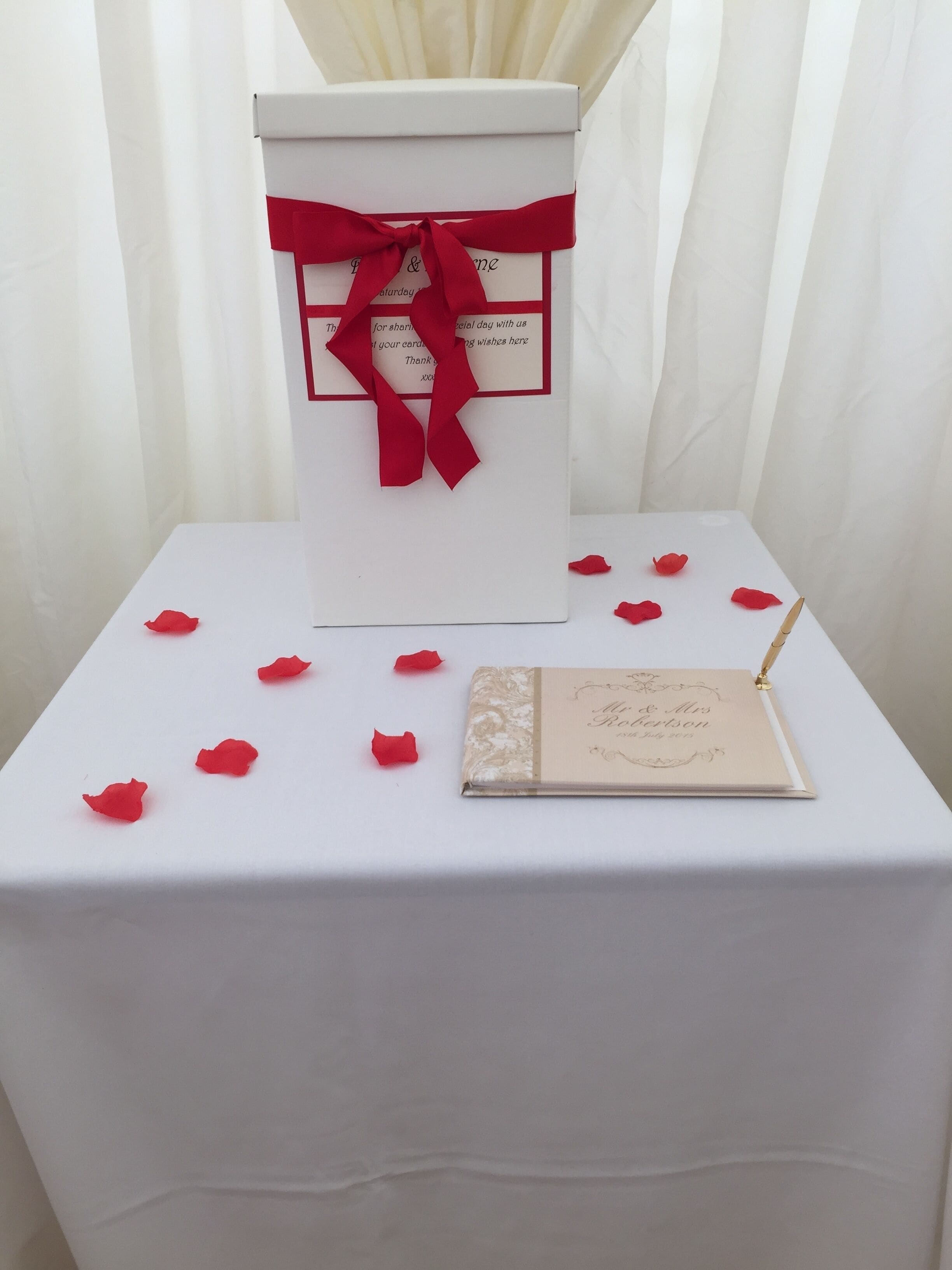 A white table with a red ribbon tied around a white box, a small booklet, and scattered red rose petals.