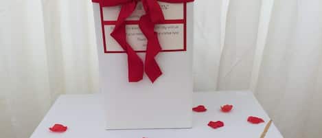 A white table with a red ribbon tied around a white box, a small booklet, and scattered red rose petals.