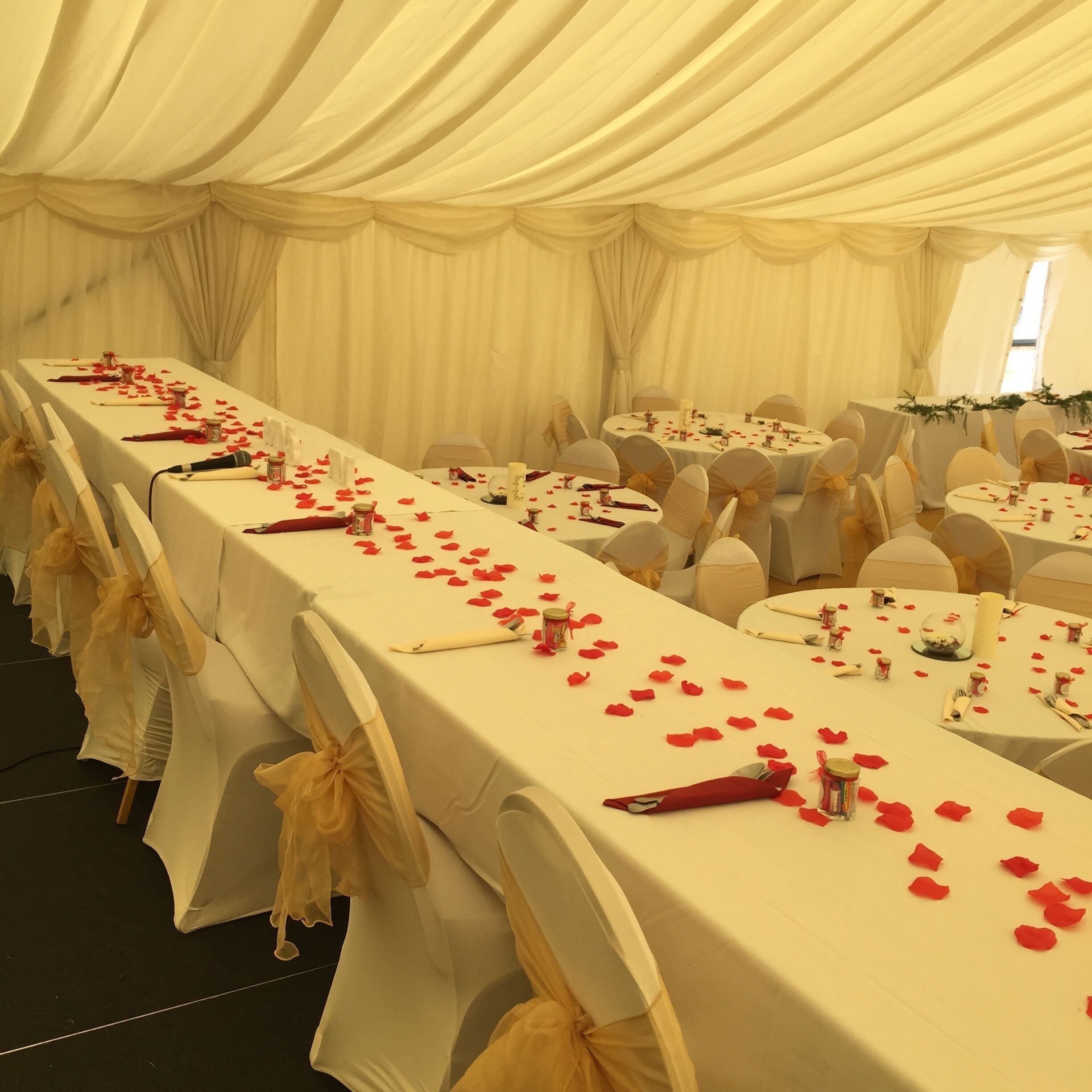 A banquet hall with long tables covered in white tablecloths, red rose petals, and candles.