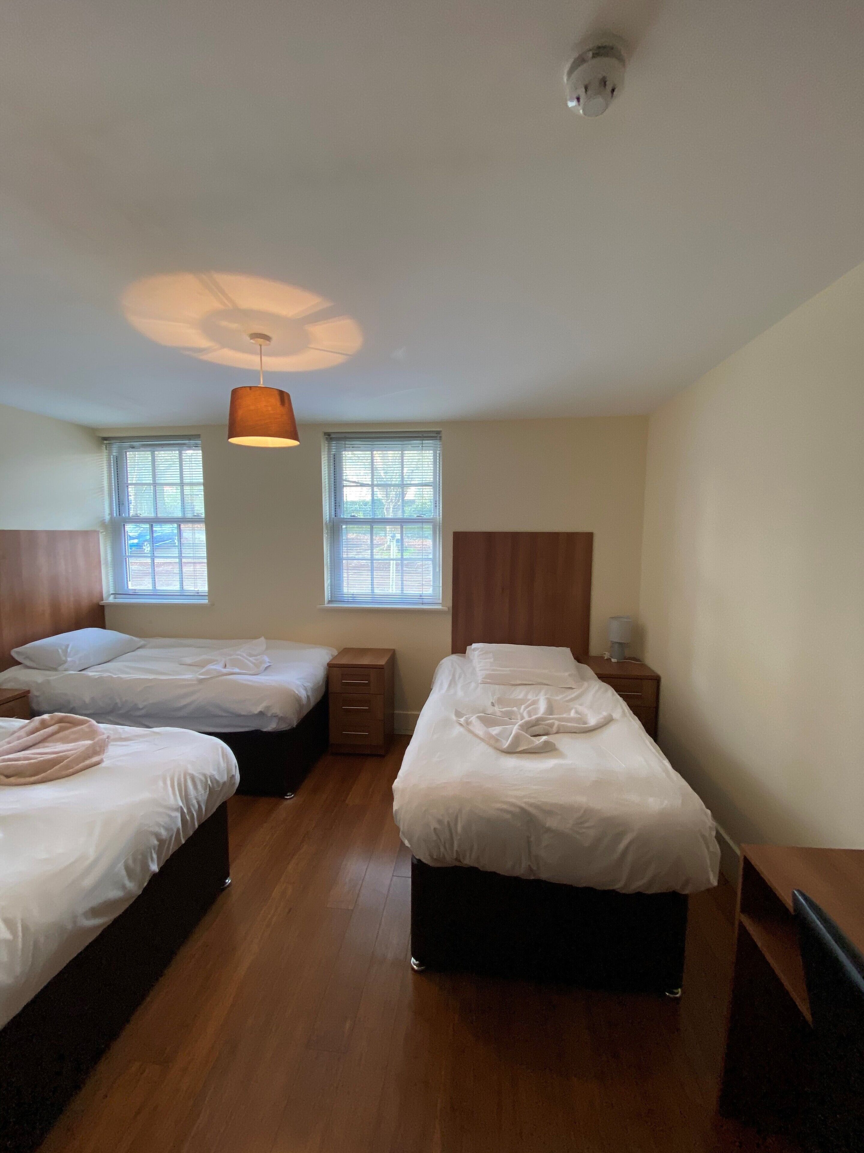 Hotellihuone, jossa on kaksi erillistä sänkyä, puulattia, kattovalaisin ja kaksi ikkunaa, joissa on sälekaihtimet.