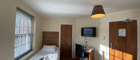 Ein kleines Hotelzimmer mit zwei Einzelbetten, einem Schreibtisch mit Computer, einem an der Wand montierten Fernseher und einem Fenster mit Jalousien.