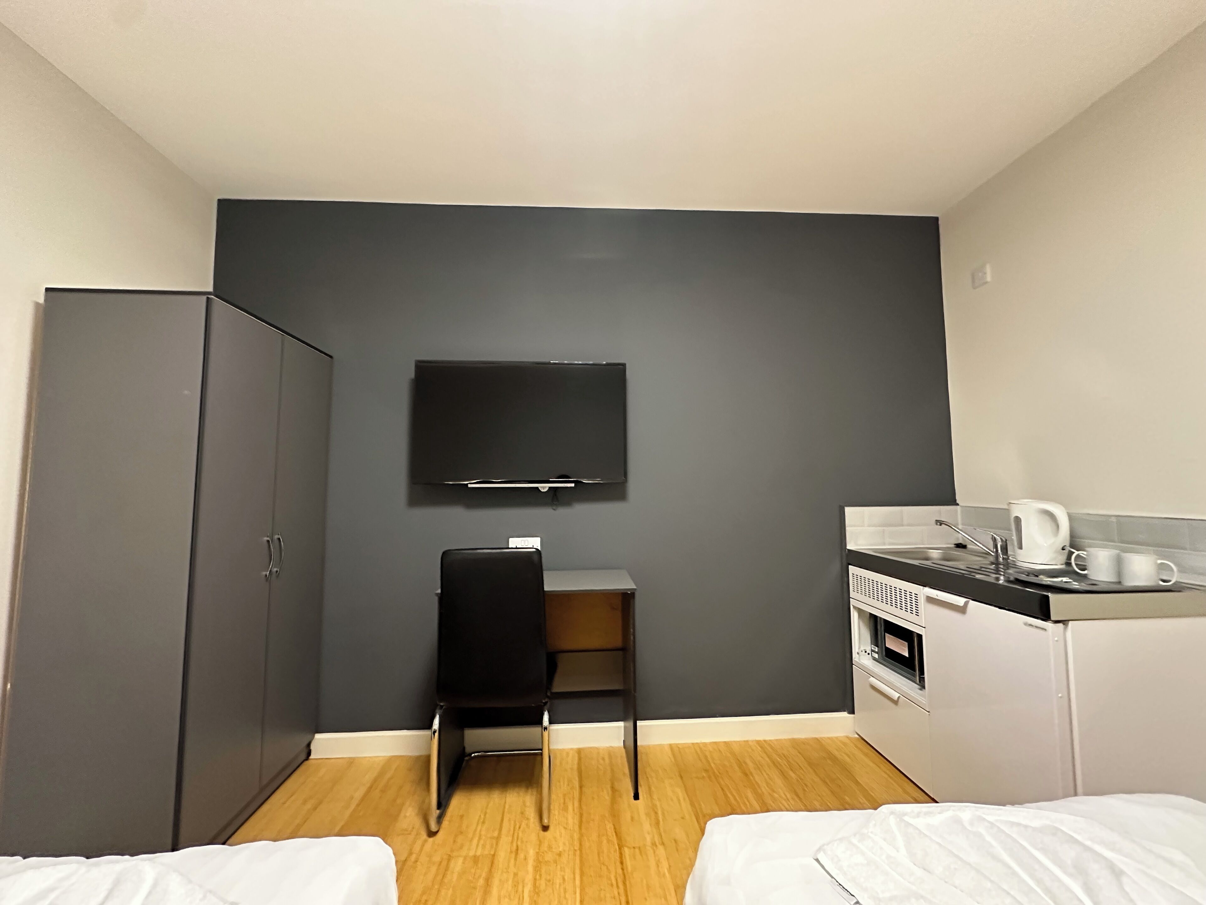 A compact hotel room with a bed, a desk with a chair, a TV mounted on the wall, a kitchenette with a coffee maker, and a wardrobe.