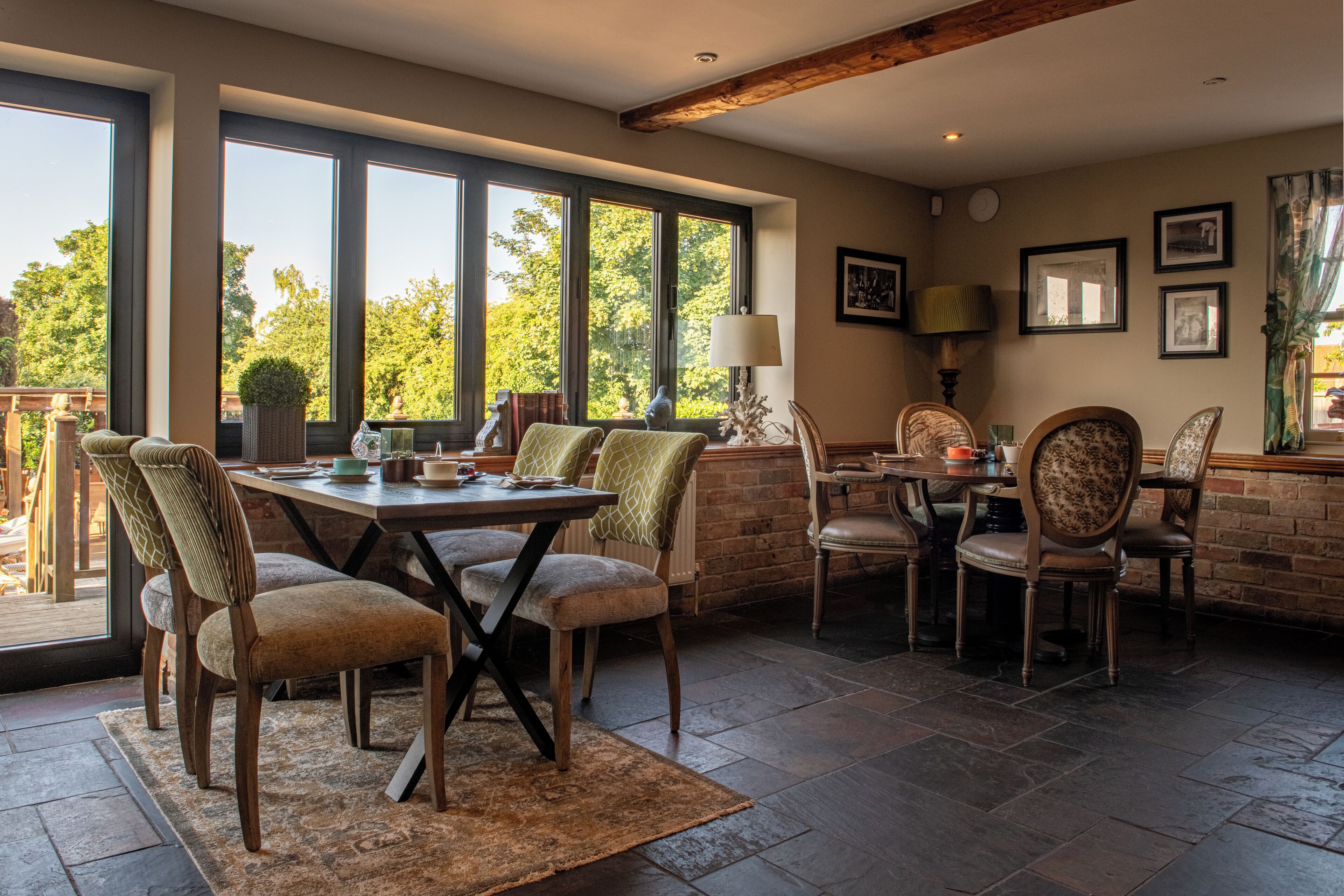 A dining area with a large table and chairs, a brick wall, and large windows with a view of trees.