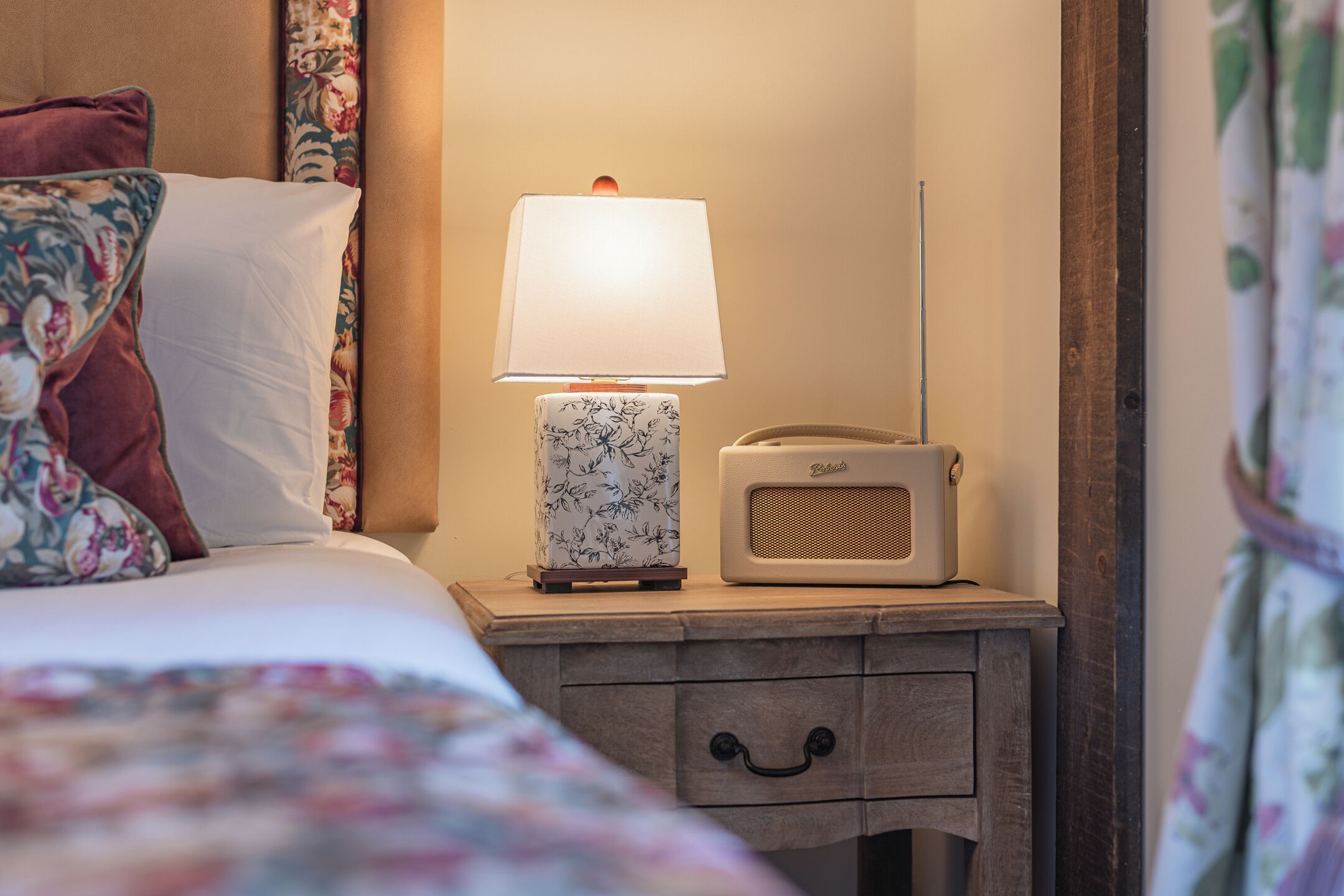 A neatly made bed with floral bedding, a bedside lamp, and a vintage radio on a wooden nightstand.