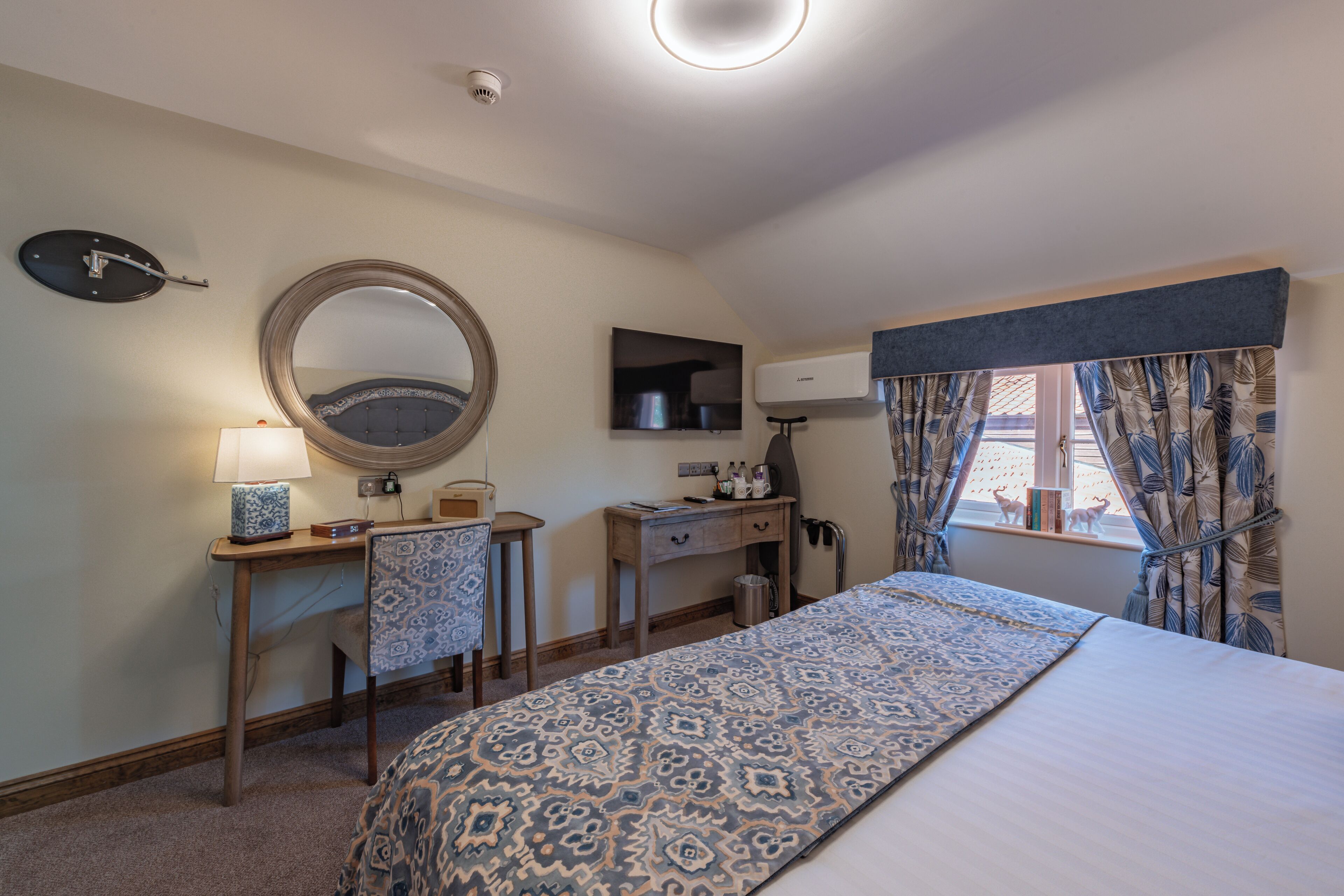 A bedroom with a bed, a desk, a chair, a mirror, a lamp, a window with curtains, and a wall-mounted heater.