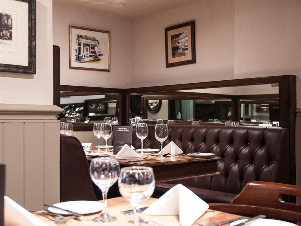 L’intérieur d  ’un restaurant présente un coin salon avec des banquettes en cuir foncé, des tables soigneusement dressées avec des verres à vin, et des tableaux encadrés accrochés aux murs.