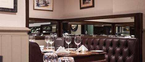 Un interior de restaurante con un banco de cuero oscuro, mesas de comedor ordenadas con copas de vino y cuadros en las paredes.