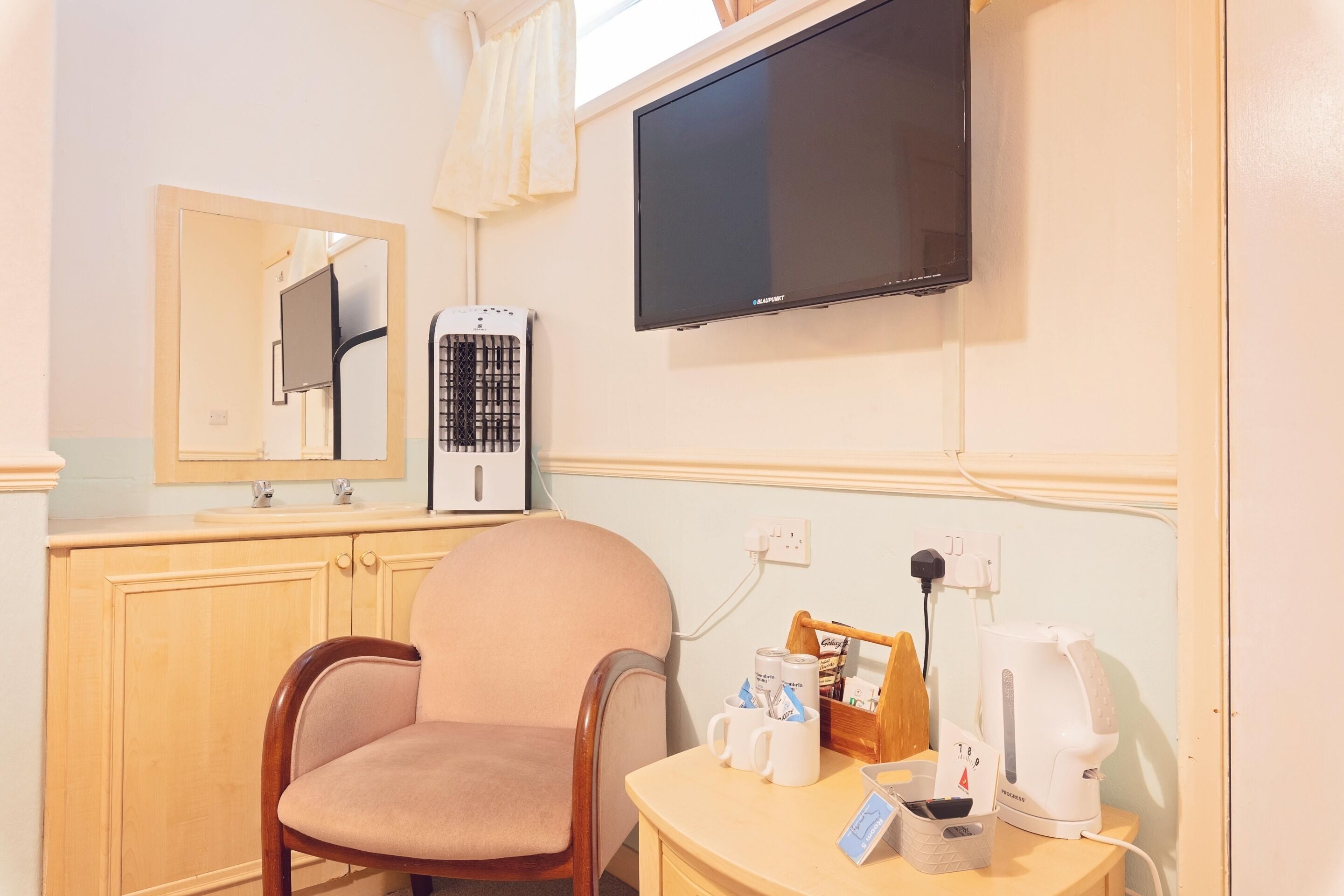 A hotel room with a wooden chair, a small table with a coffee maker, a television, and a heater.