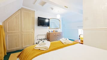A hotel room with a bed, a TV mounted on the wall, a wooden wardrobe, a mirror, and a bathroom with a sink and towel rack.