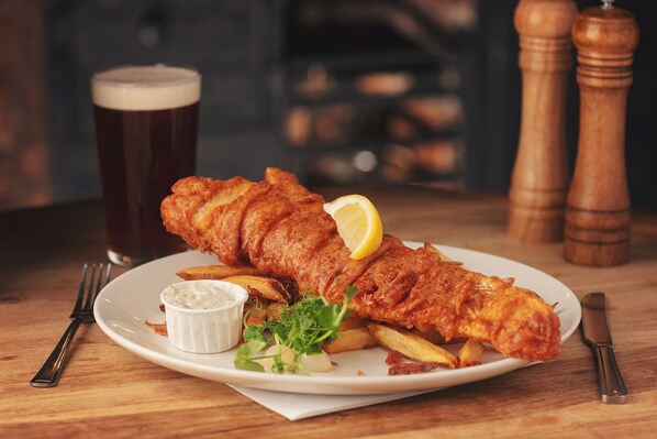 A plate of fish and chips with a glass of beer, garnished with a lemon slice, on a wooden table. - The Tavern - Steakhouse and Lodge (Alnwick)