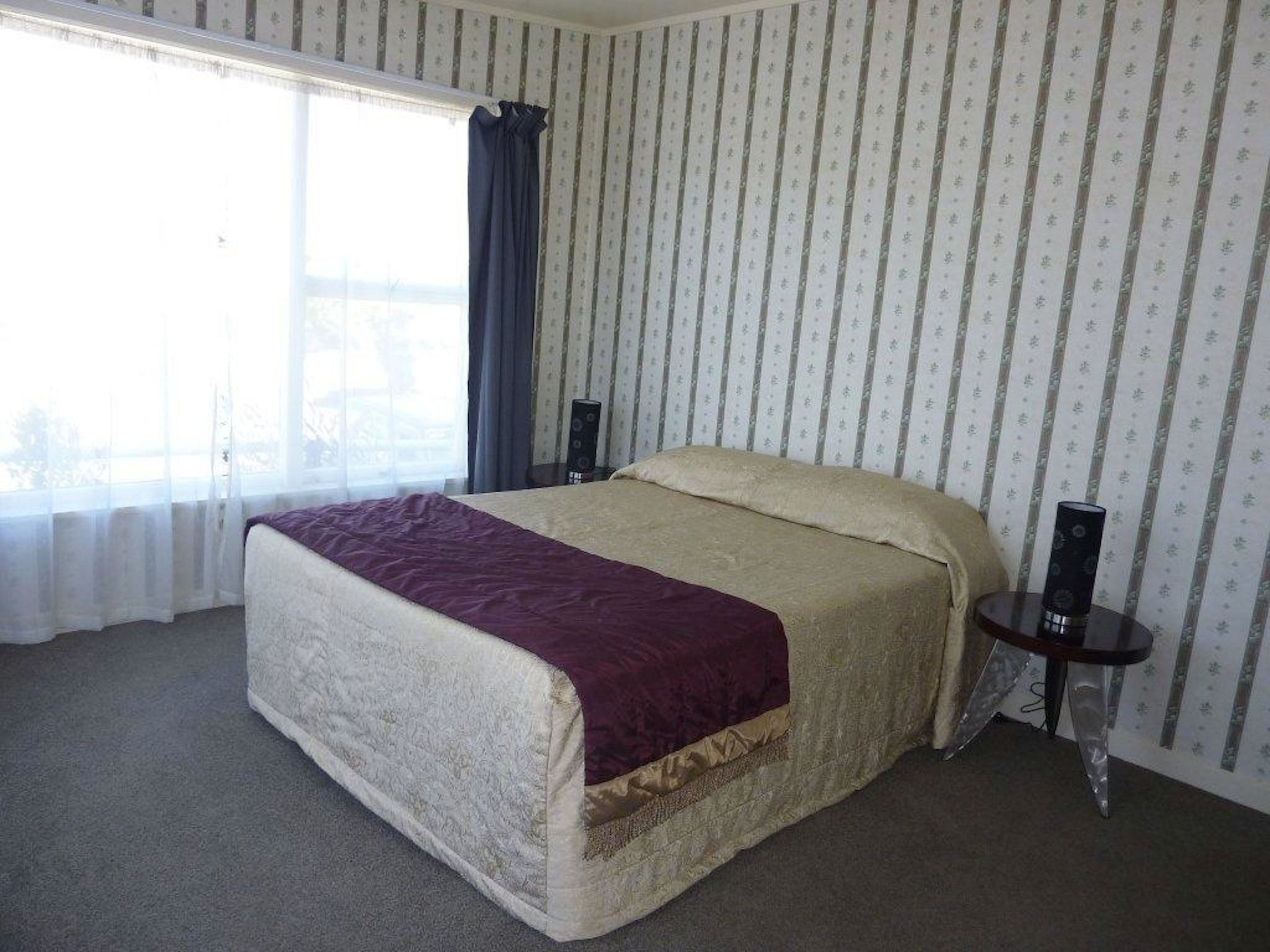 A bedroom with a large bed, a bedside table, a window with curtains, and a patterned wallpaper.