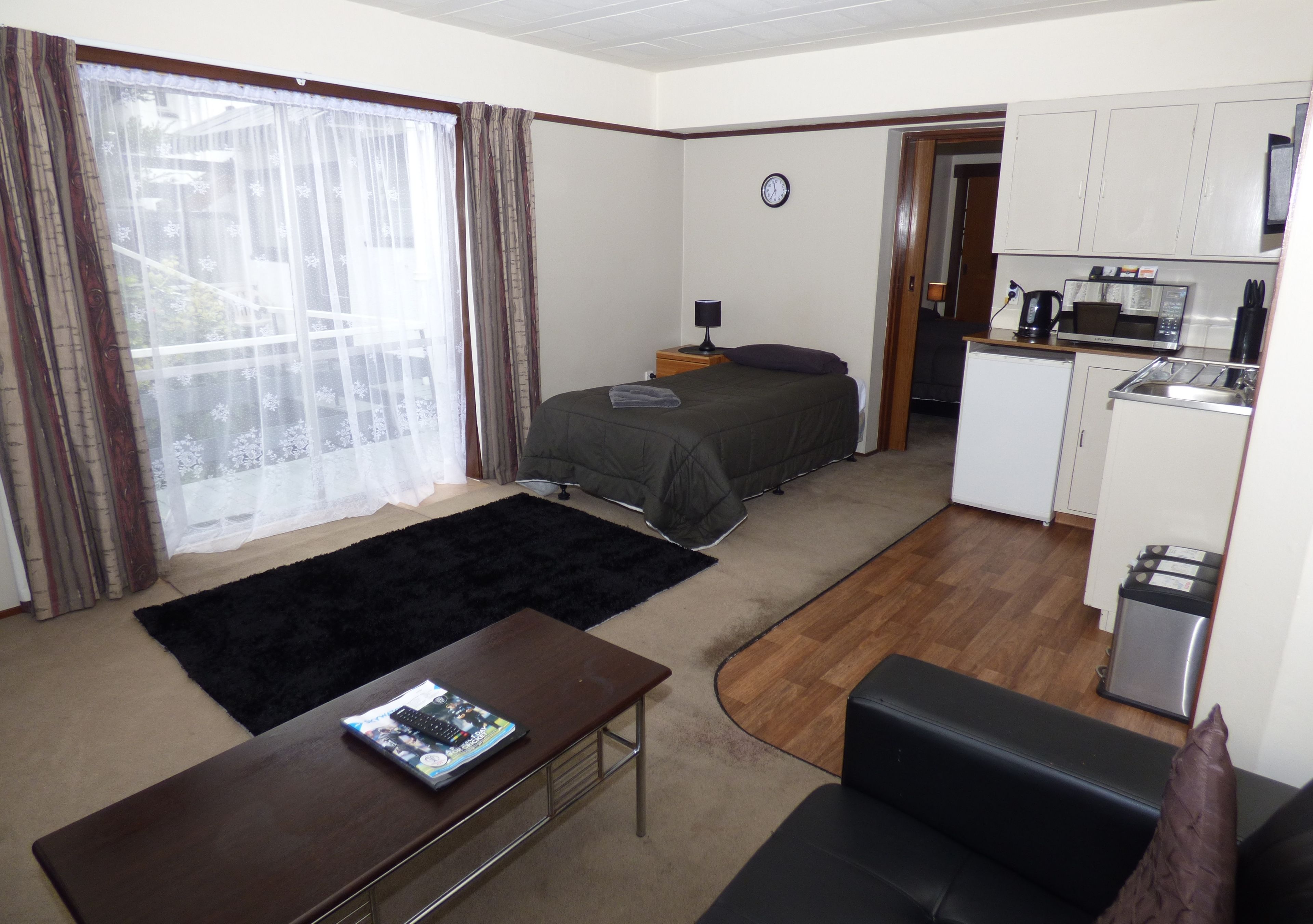 Habitación de hotel pequeña con cama, un escritorio, kitchenette y vistas a través de una ventana con cortinas.