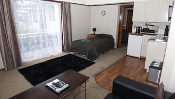 Habitación de hotel pequeña con cama, un escritorio, kitchenette y vistas a través de una ventana con cortinas.