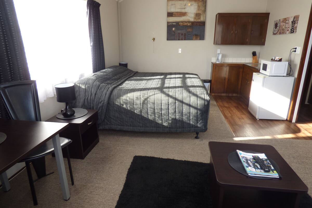 A compact hotel room with a bed, a desk with a chair, a small kitchenette, and a window with curtains.