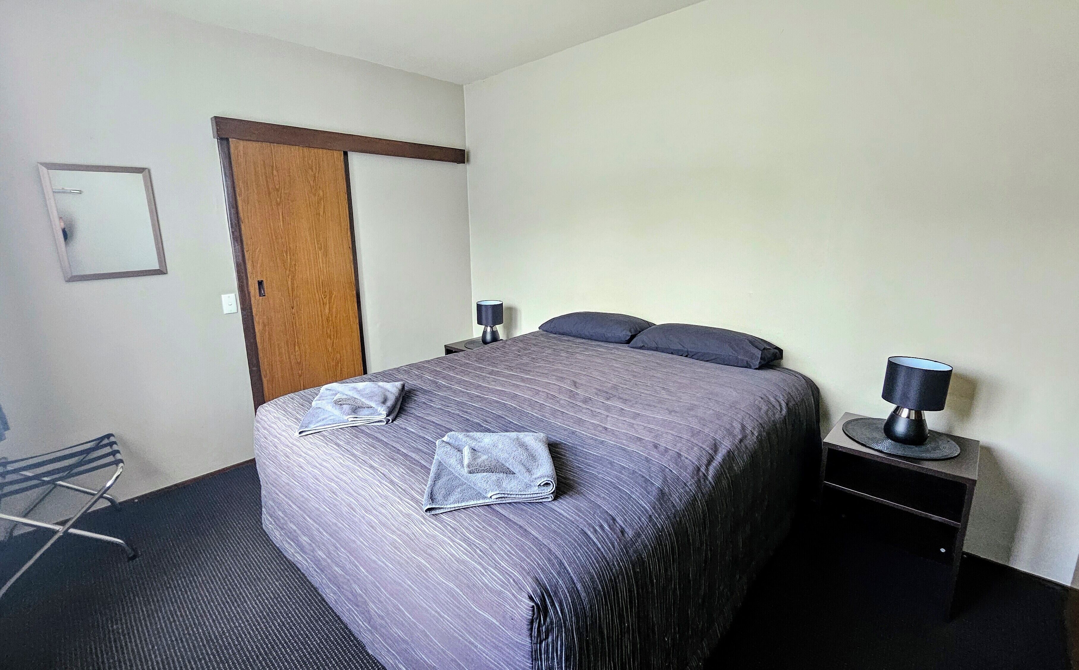 A neatly made bed with a grey bedspread and two pillows, a wooden door, a small bedside table with a lamp, and a chair with a metal frame.