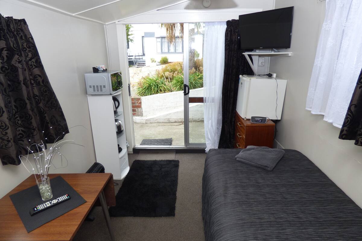 A small, modern room with a bed, a desk with a computer, a TV, and a window with curtains.
