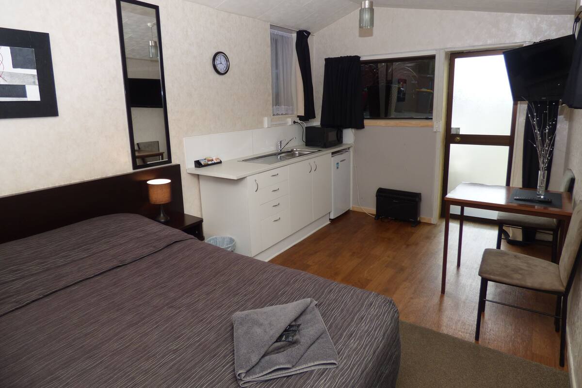 A compact hotel room with a bed, a small kitchenette, a desk, and a window.
