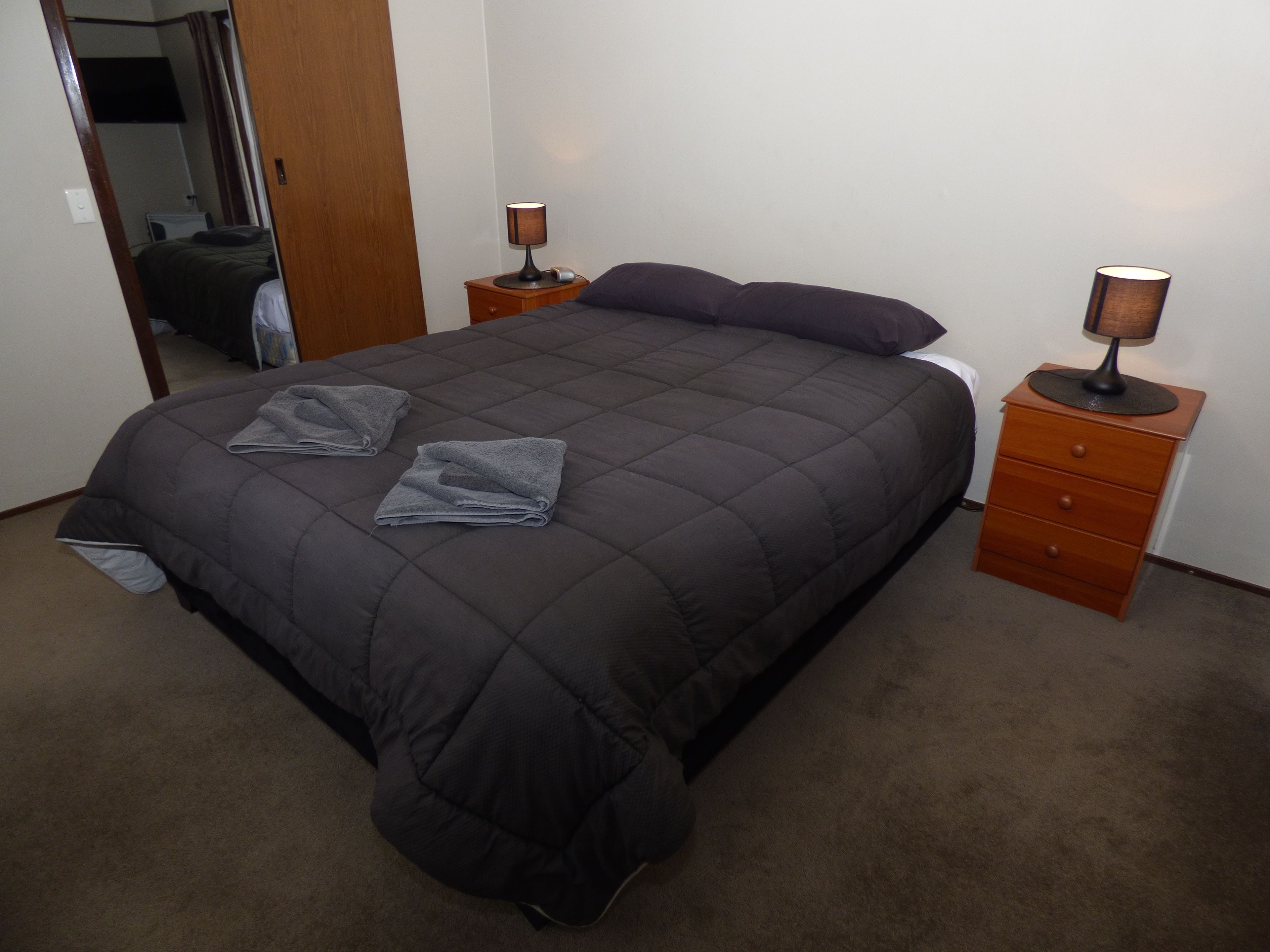 Cama bem arrumada com colcha escura, dois travesseiros e duas toalhas dobradas. Há duas mesas de cabeceira com abajures e um móvel de madeira com gavetas.
