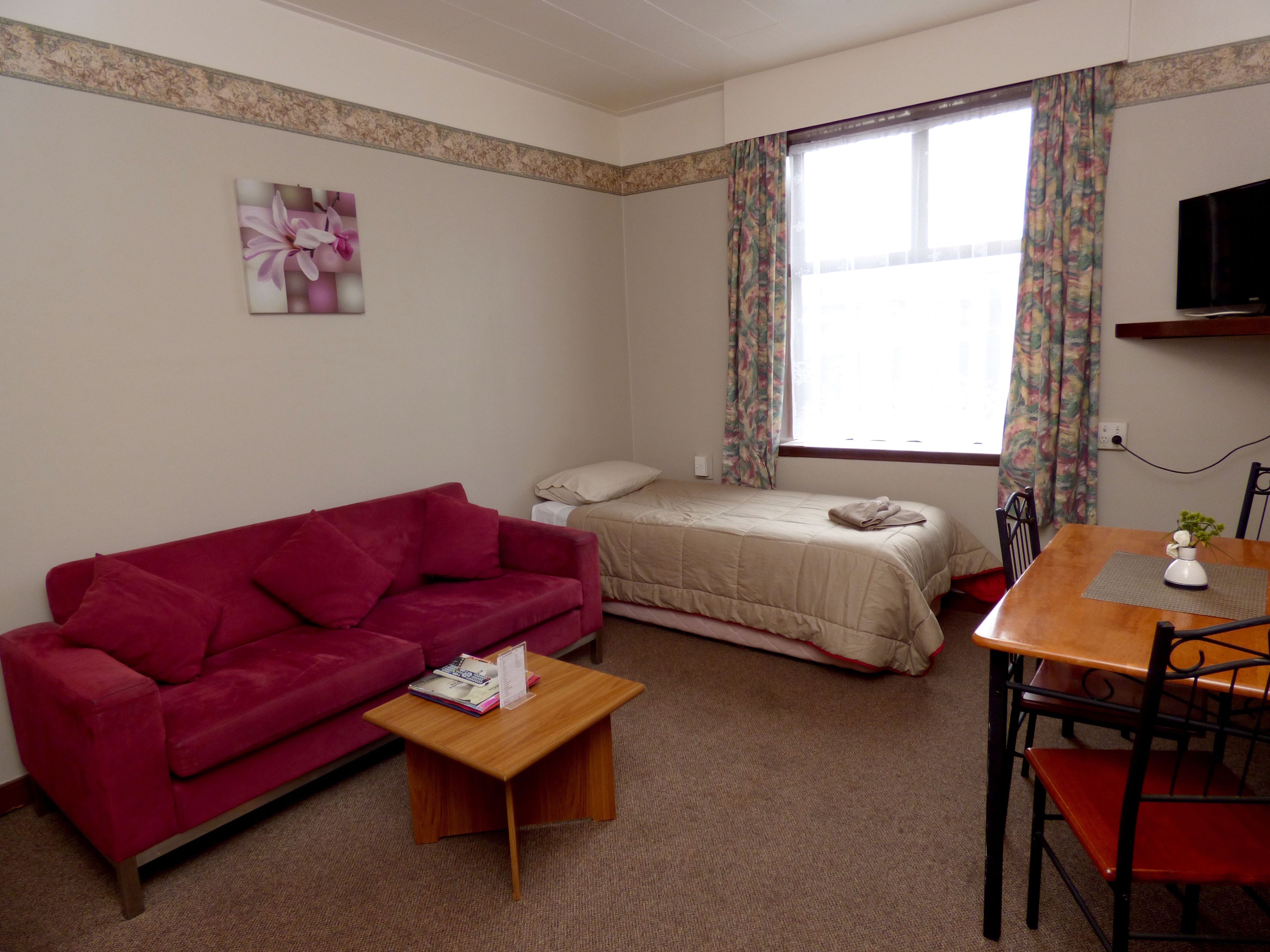 Una habitación compacta con un sofá rojo, una pequeña mesa de madera, una cama con edredón color beige, una mesa de comedor con sillas y un televisor montado en la pared.