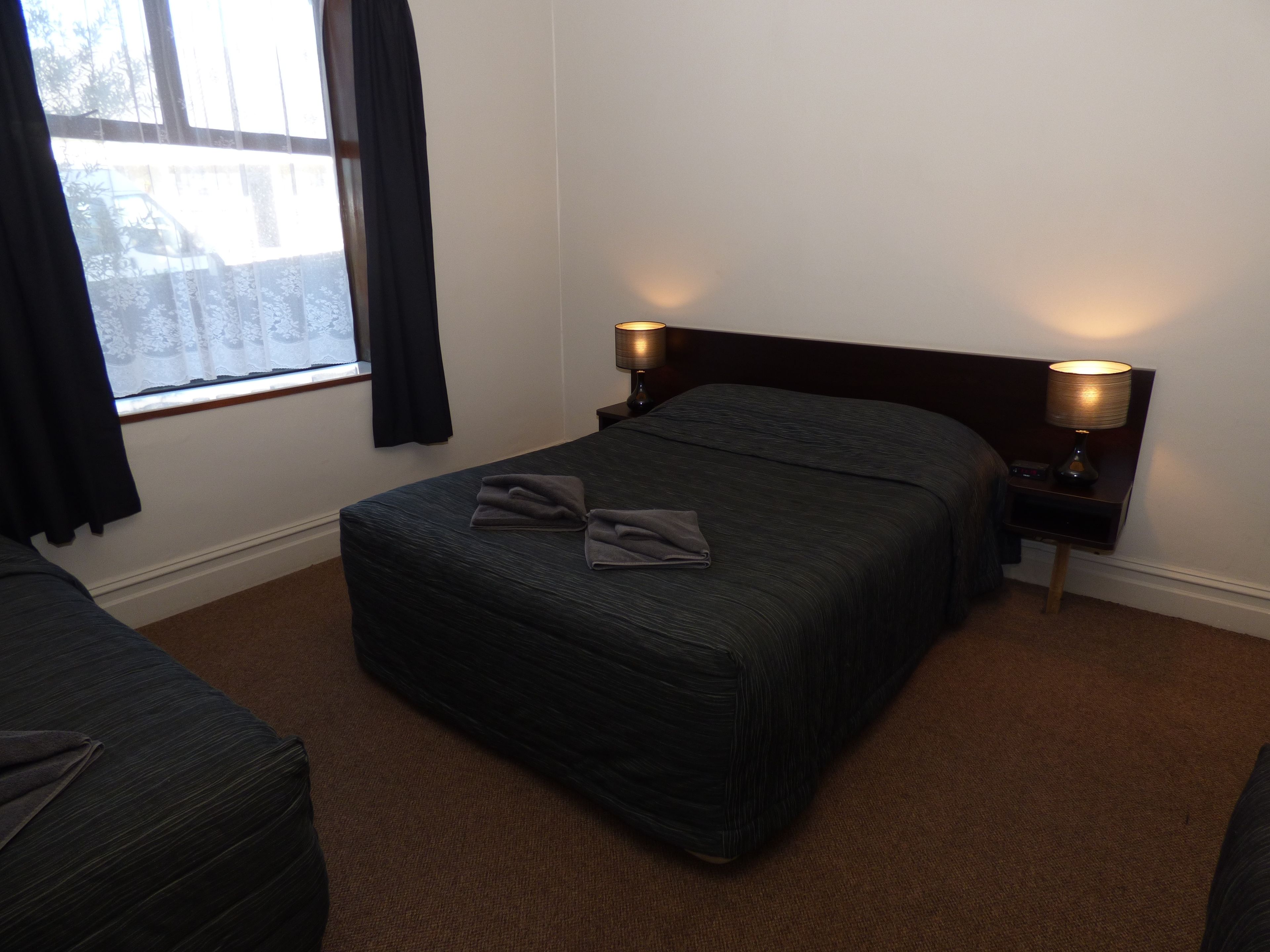 A hotel room with a bed, two bedside lamps, a window with curtains, and a carpeted floor.