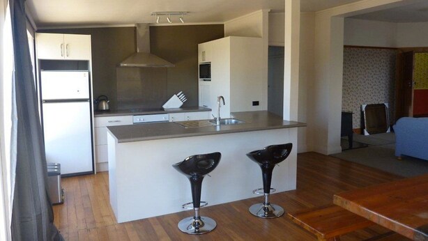 A compact kitchen with a central island, a sink, and a stove. There are two bar stools and a dining table. - Anchor Motel (Timaru)
