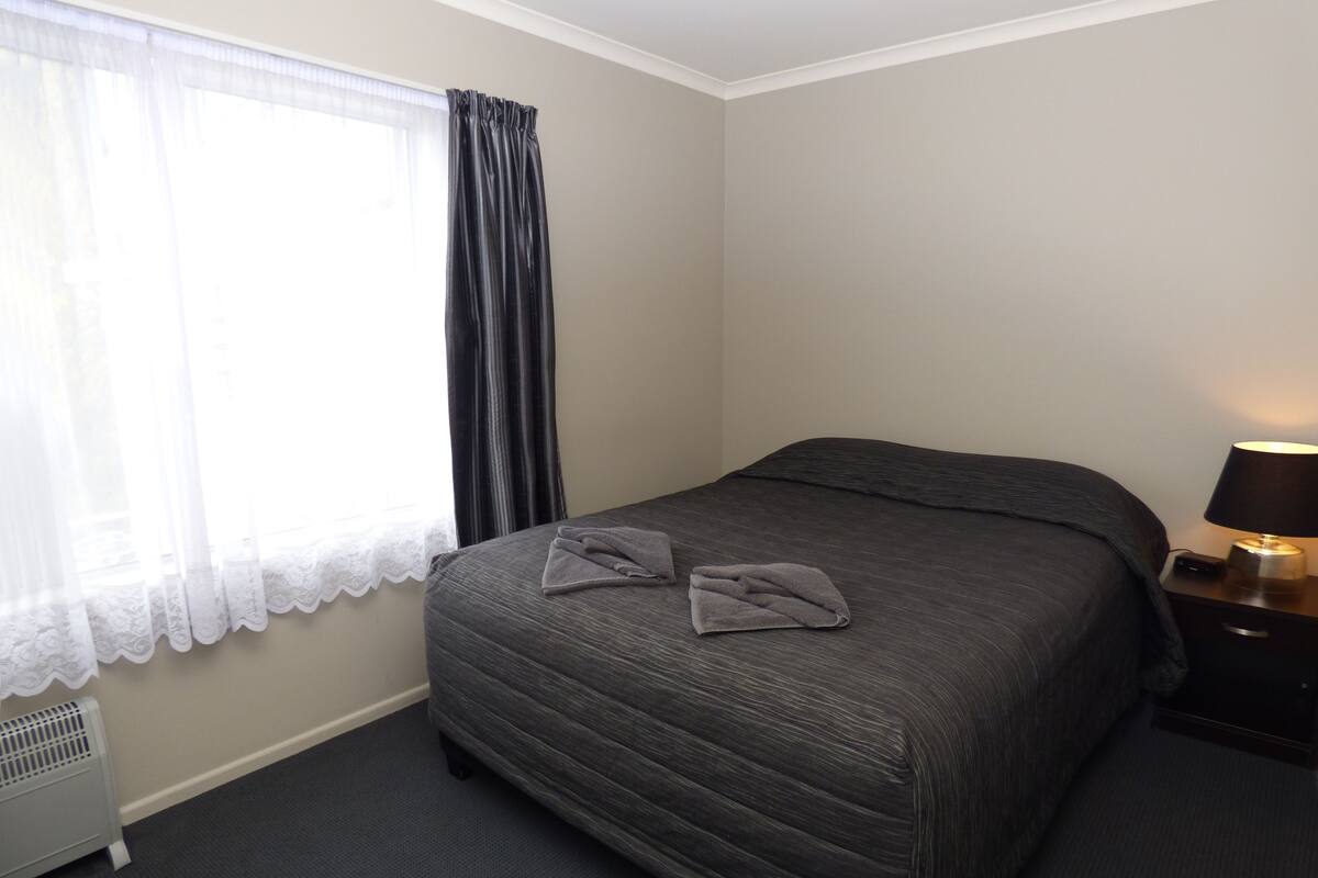 A hotel room with a bed, a nightstand with a lamp, a radiator, and a window with curtains.