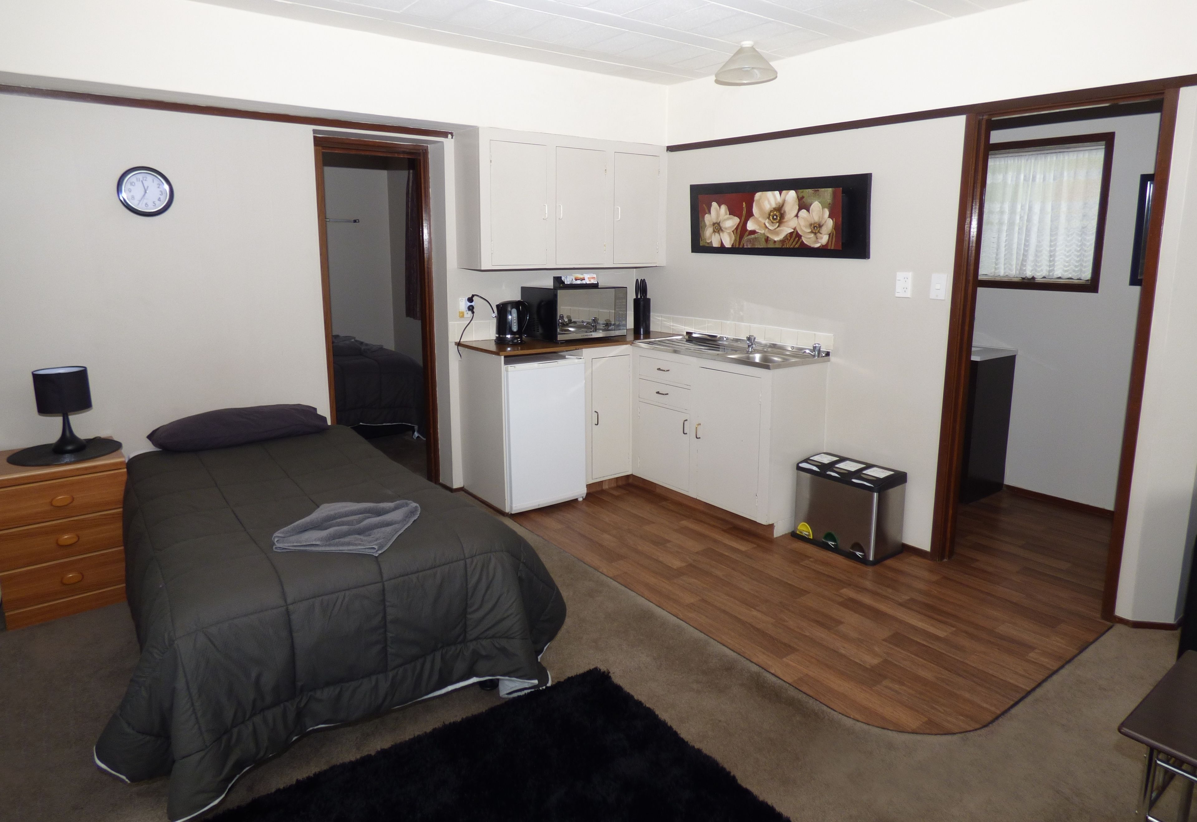 A compact room with a bed, a small kitchenette, a clock, and a door leading to another room.