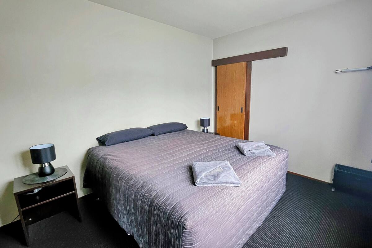 A neatly made bed with a grey bedspread and pillows, a bedside table with a lamp, and a wooden wardrobe.