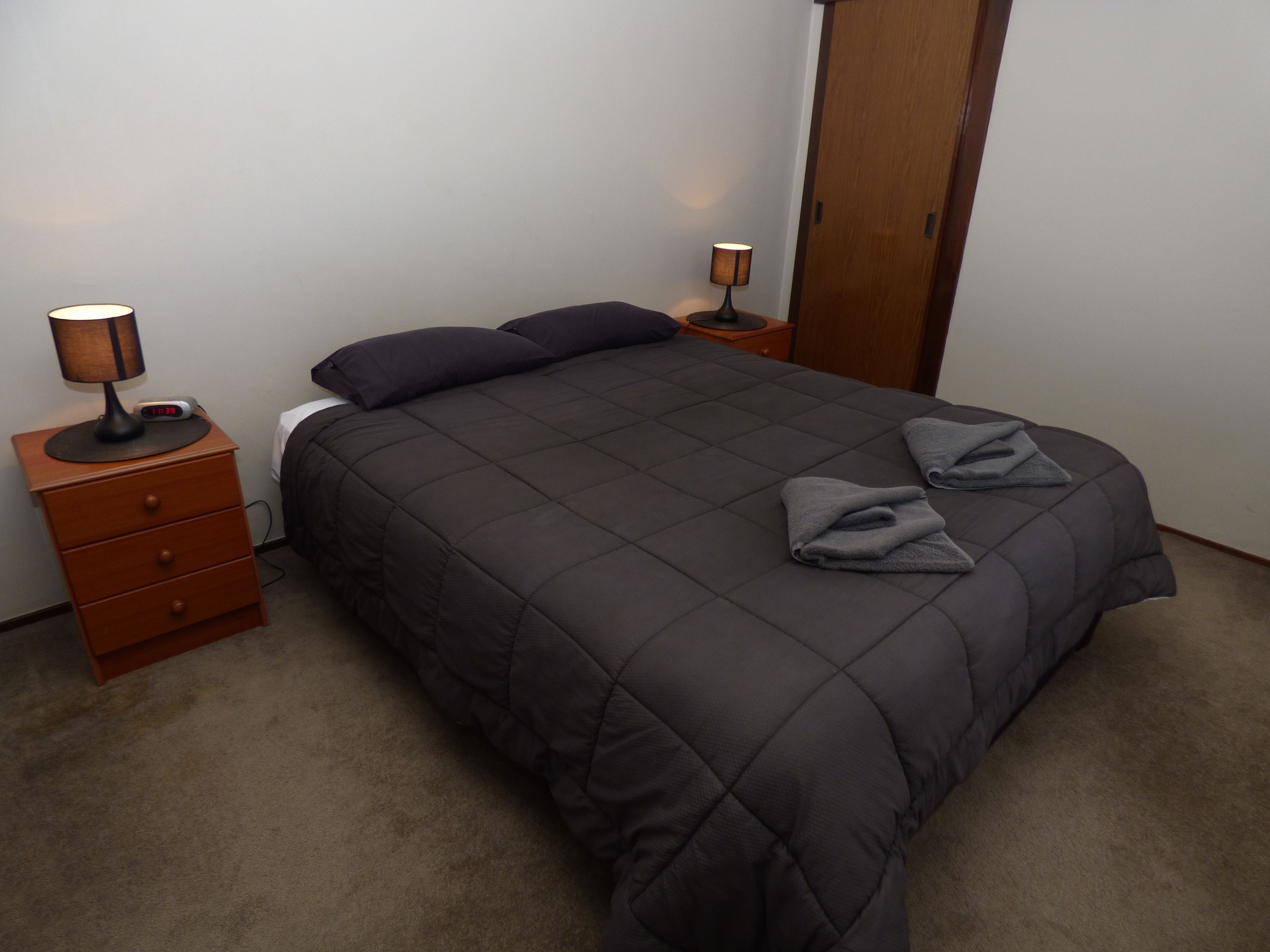 Ein ordentlich gemachtes Bett mit einer dunklen Decke, zwei Kissen und zwei gefalteten Handtüchern. Ein Holznachttisch mit Lampe und digitaler Uhr steht daneben. Der Raum hat eine Holztür und eine schlichte weiße Wand.