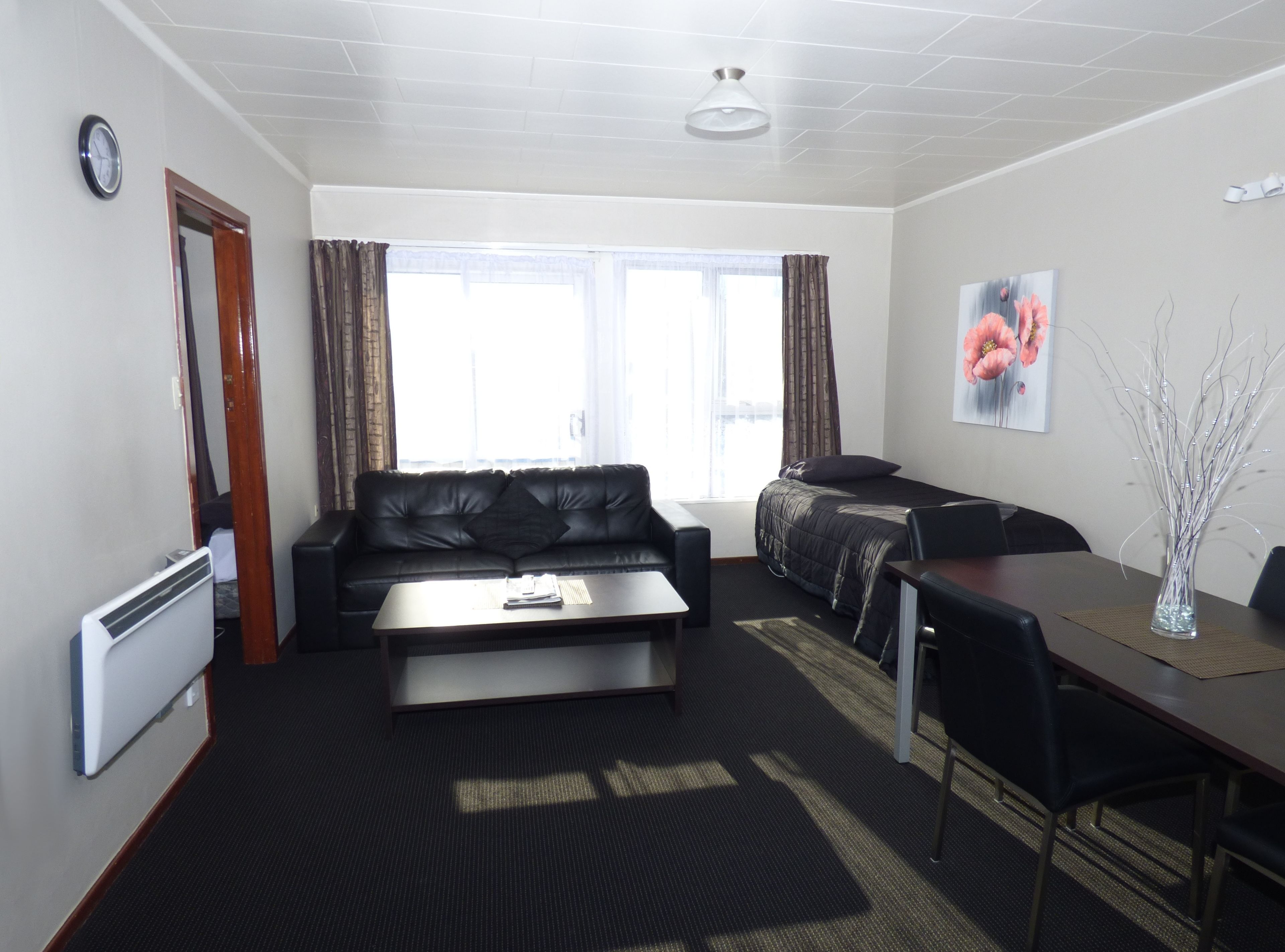 A modern hotel room with a black leather sofa, a small dining table, a bed with a dark duvet, a large window with curtains, a clock on the wall, and a painting of flowers.