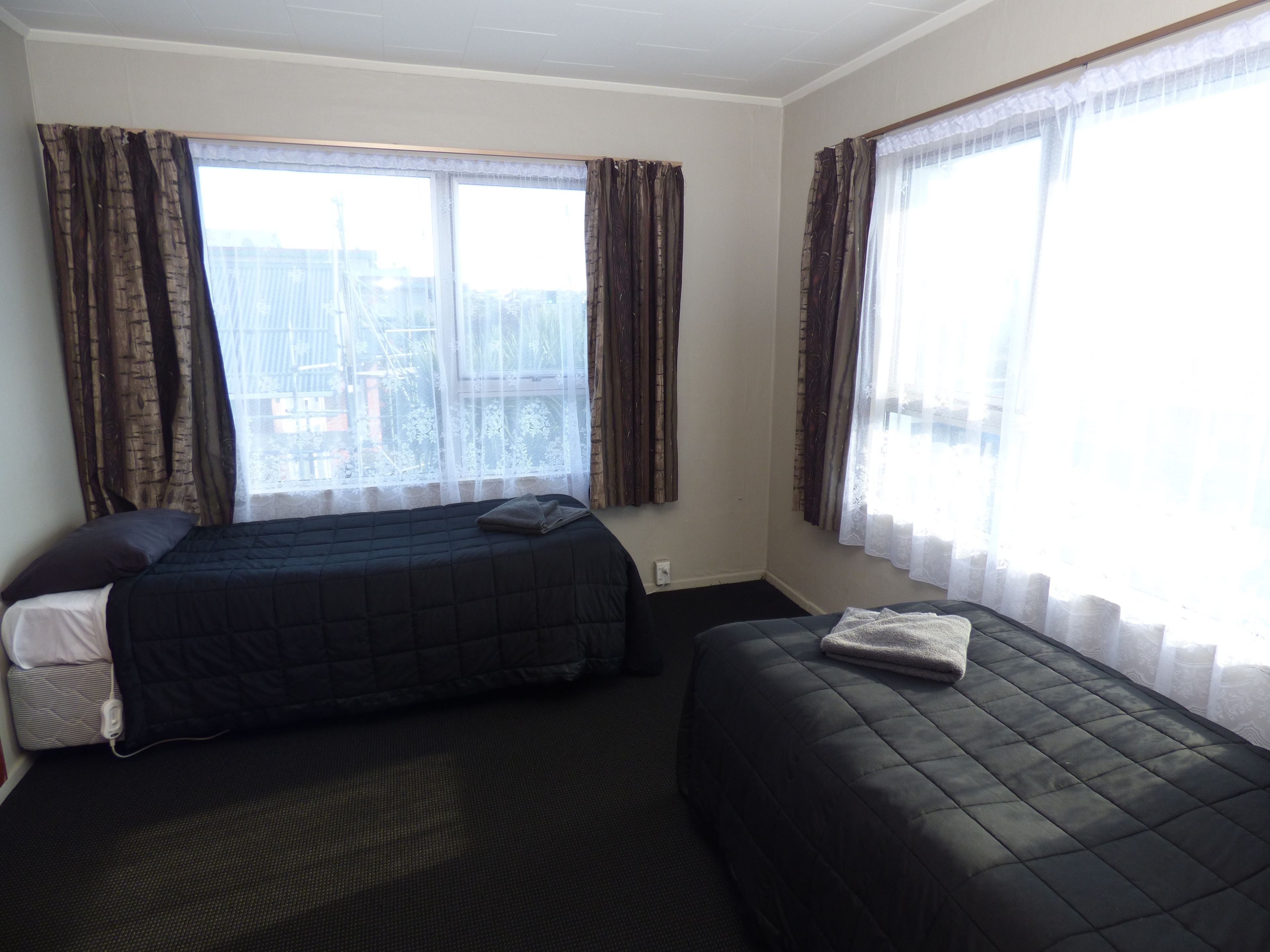 A hotel room with two beds, a window with curtains, a small bedside table, and a power outlet.