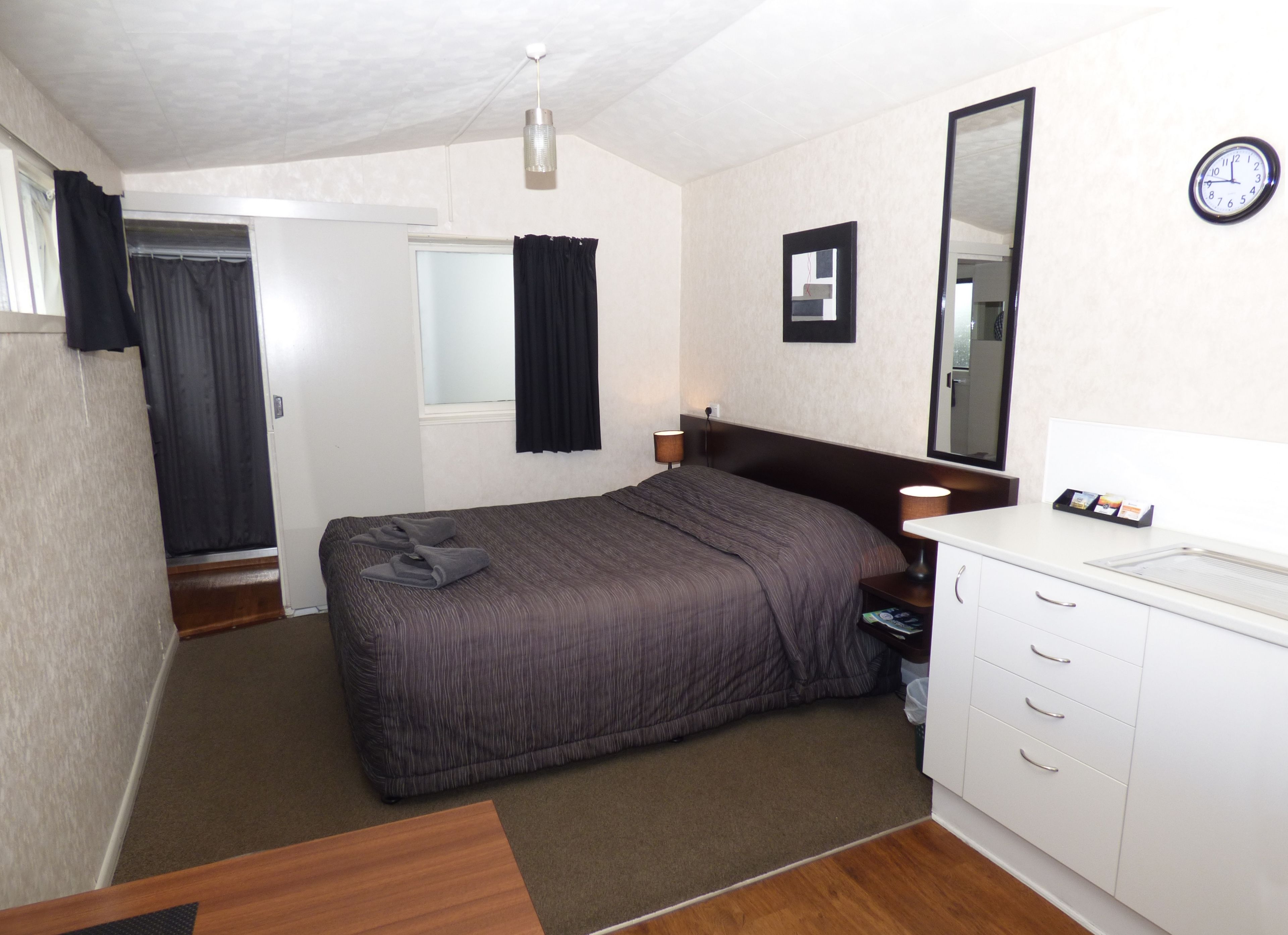 A hotel room with a bed, a bedside table with a lamp, a mirror, a clock, and a small desk with drawers.