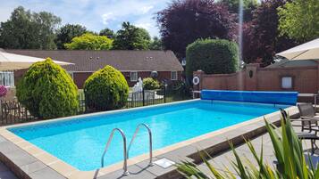 A rectangular swimming pool with a tiled edge, surrounded by a low brick wall. There are two metal pool ladders, a blue pool cover, and a small garden area with a hedge and a statue.