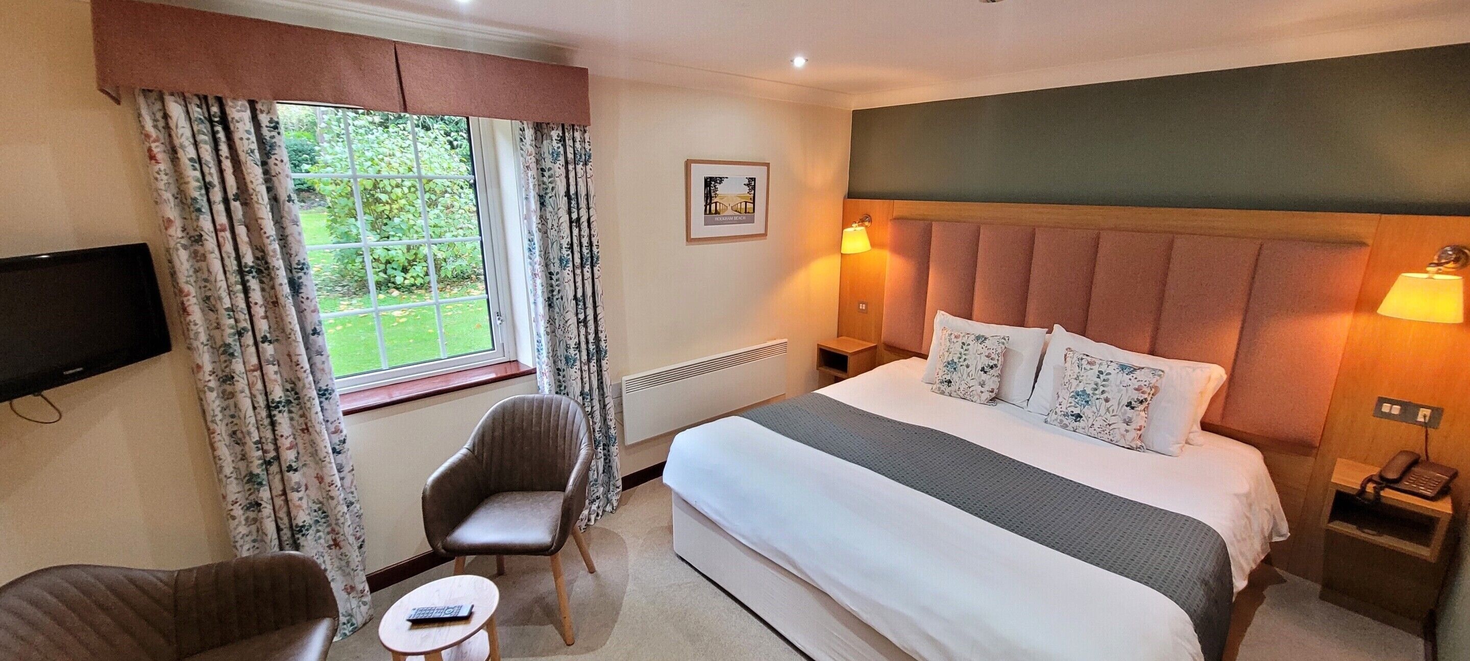 A hotel room with a large bed, a television mounted on the wall, a chair, a small table, and a window with curtains.