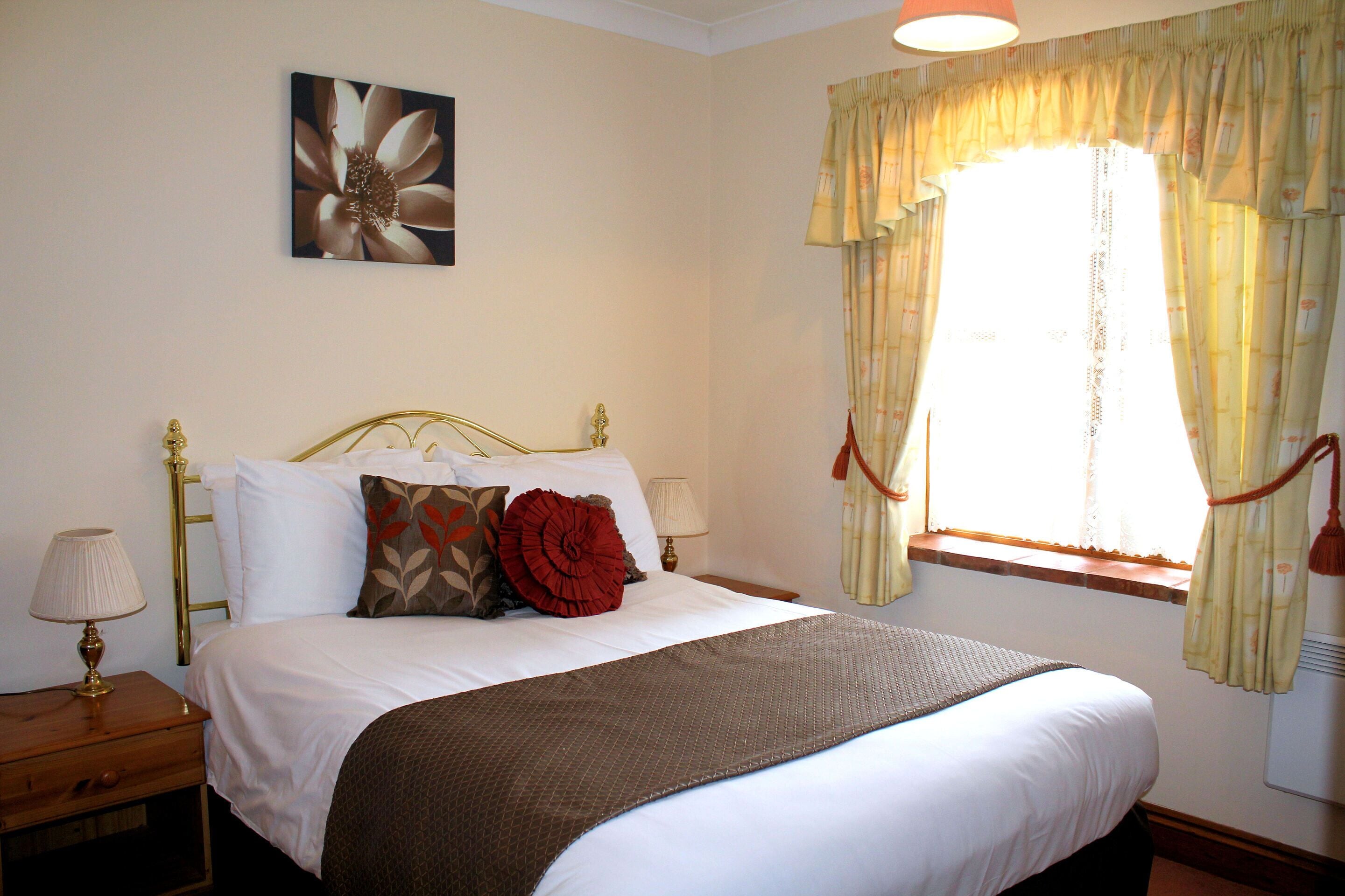 A neatly made bed with white linens and a brown throw, a nightstand with a lamp, and a window with floral curtains.