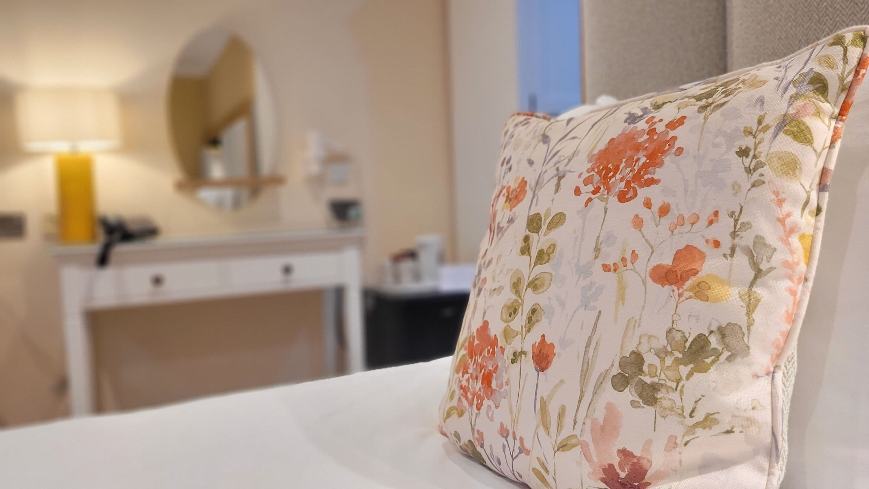 Et hotelværelse med et blomstret pude på sengen, en hvid kommode, et spejl og en lampe.